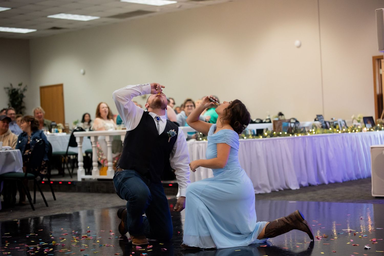 Couple kneeling on dance floor with confetti at wedding reception celebration.