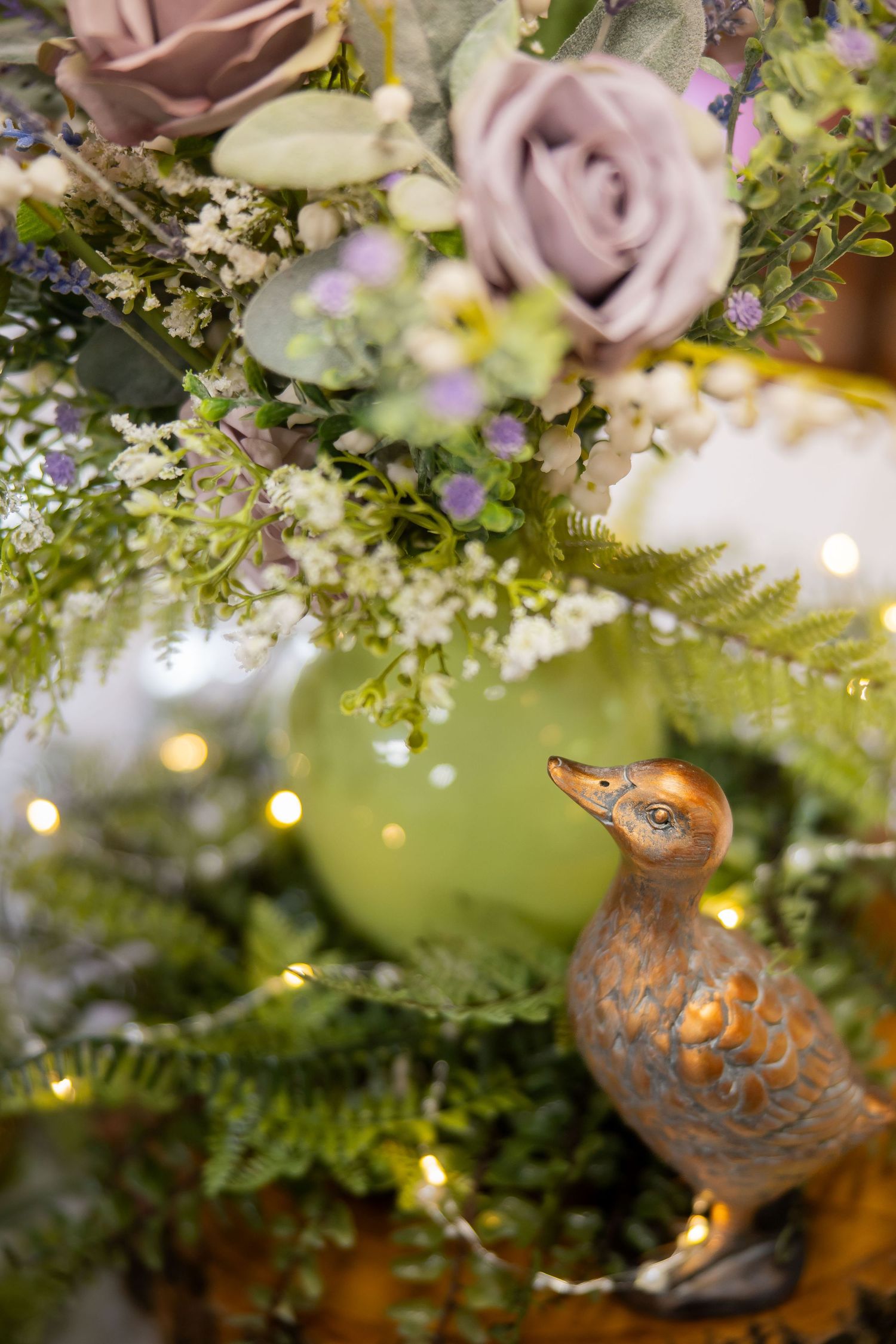 Floral wedding centerpiece with purple roses, ferns, fairy lights, and a decorative duck figurine.