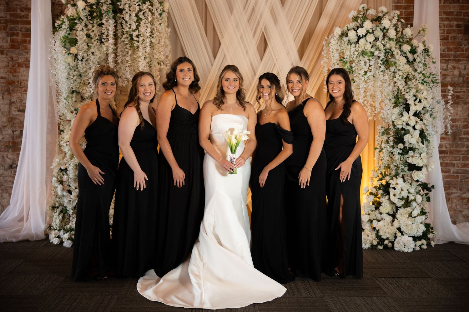 Bride in white gown with six bridesmaids in black dresses posing before floral backdrop at elegant wedding.