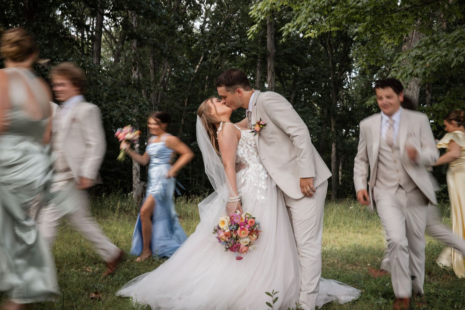 Bride and groom share a romantic kiss surrounded by blurred wedding party in a lush outdoor garden setting.