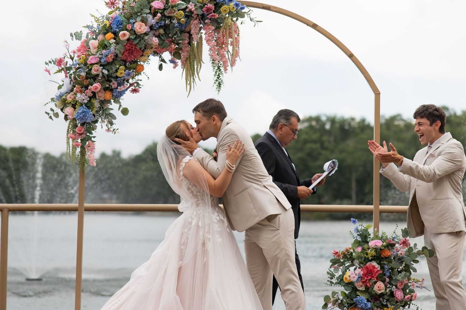 Bride and groom share first kiss under floral arch with colorful blooms at outdoor lakeside wedding ceremony.