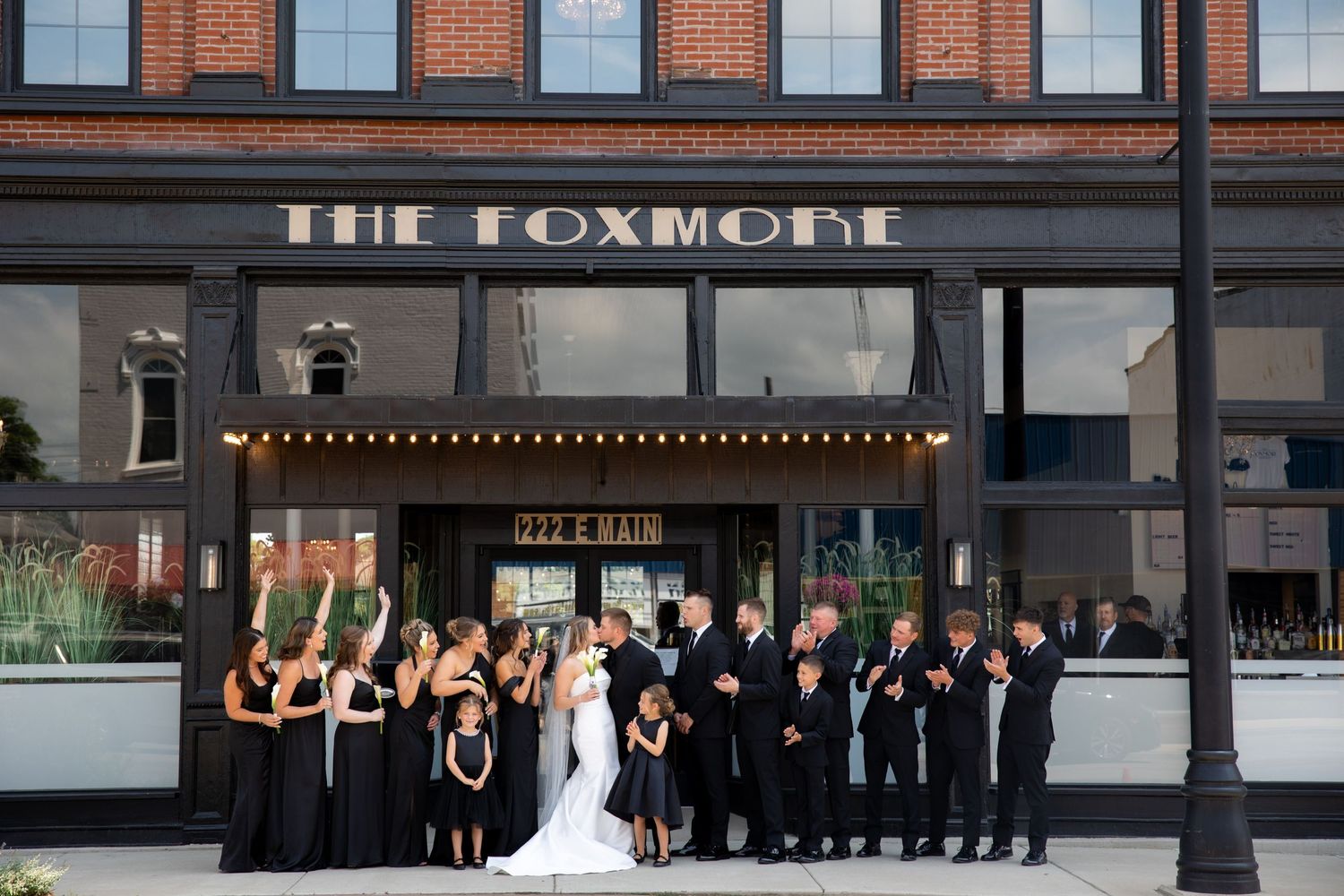 Wedding party poses in formal attire outside The Foxmore venue with illuminated marquee signage.