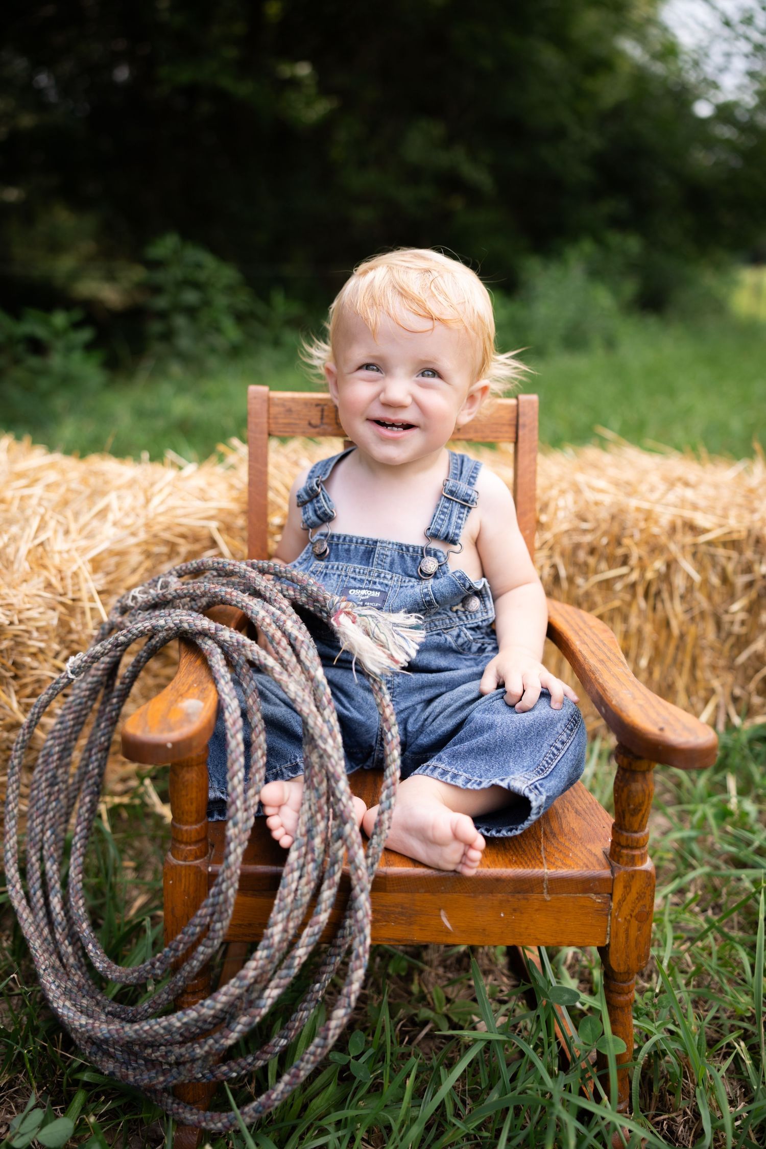 A smiling toddler in denim overalls sits in a wooden chair next to a coiled rope on hay bales.