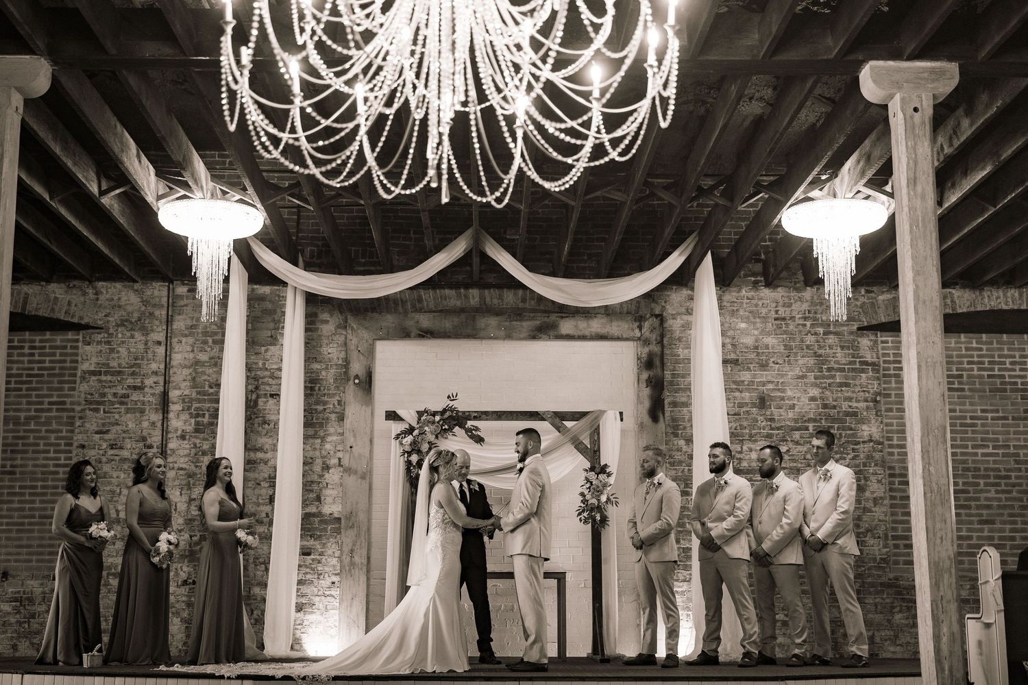 Black and white wedding ceremony photo with elegant chandelier and draped fabric in rustic brick venue.