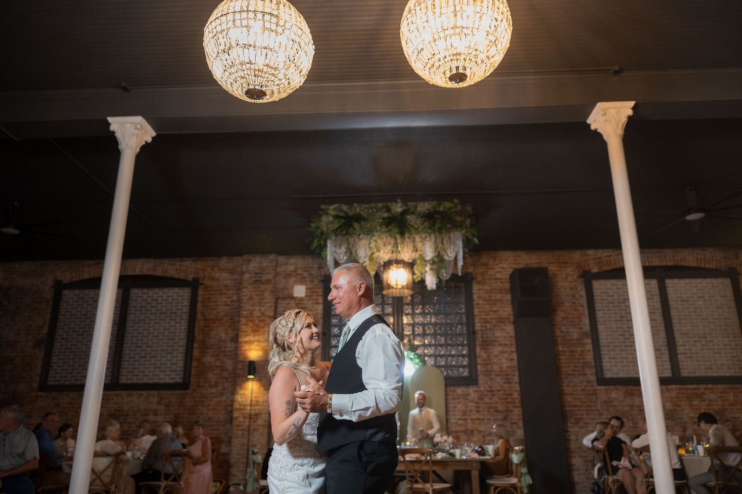 Couples dance together beneath crystal chandeliers during an elegant wedding reception in a rustic brick venue.