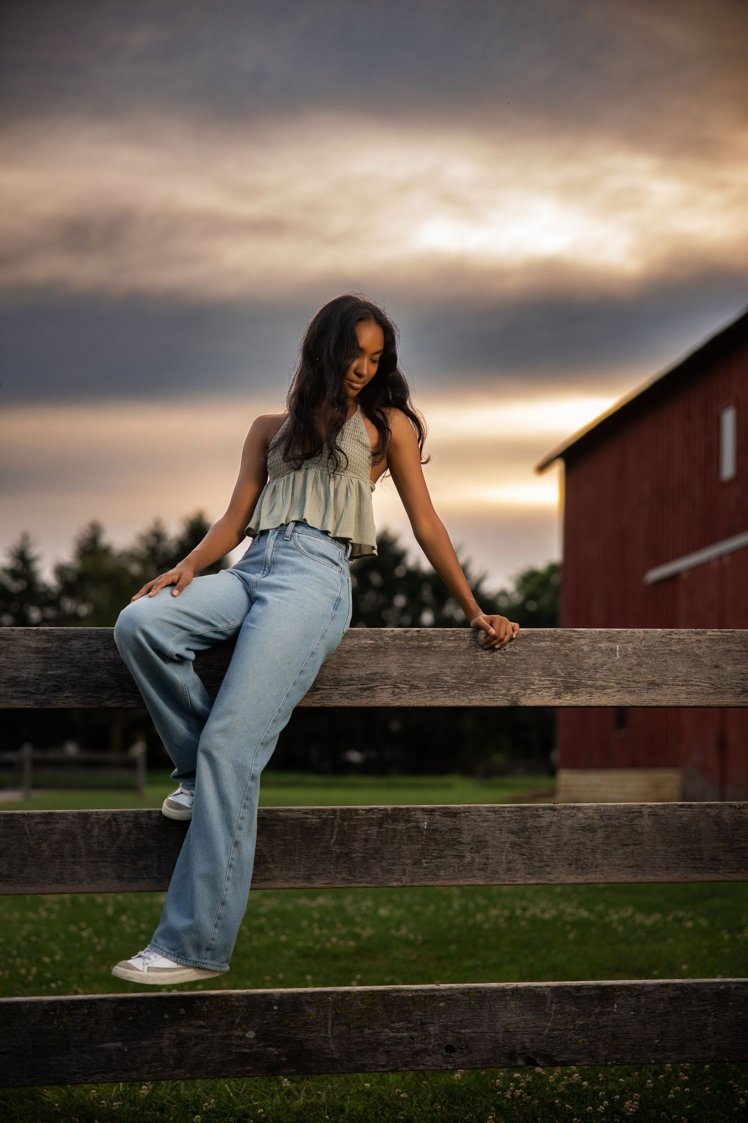 A person in casual denim sits on wooden fence rails during golden hour with a red barn in background.