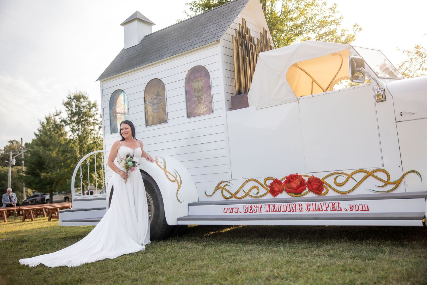 A wedding dress train flows elegantly across green grass outside a white chapel on a sunny day.