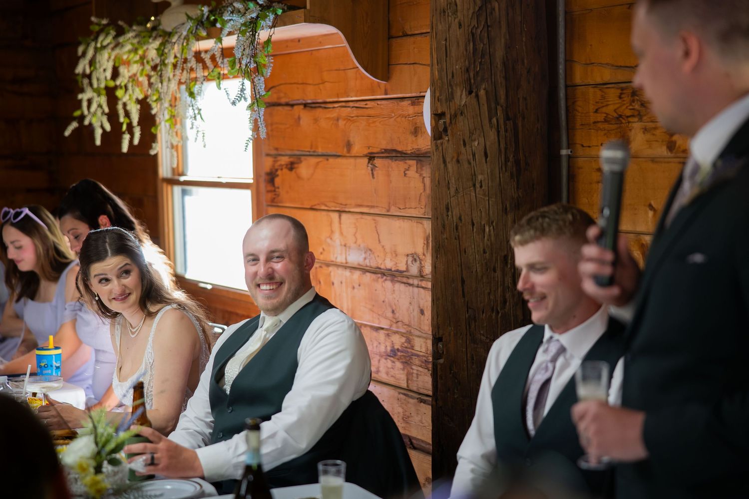 Groom in vest laughs during wedding reception speech inside a warm rustic log cabin venue.