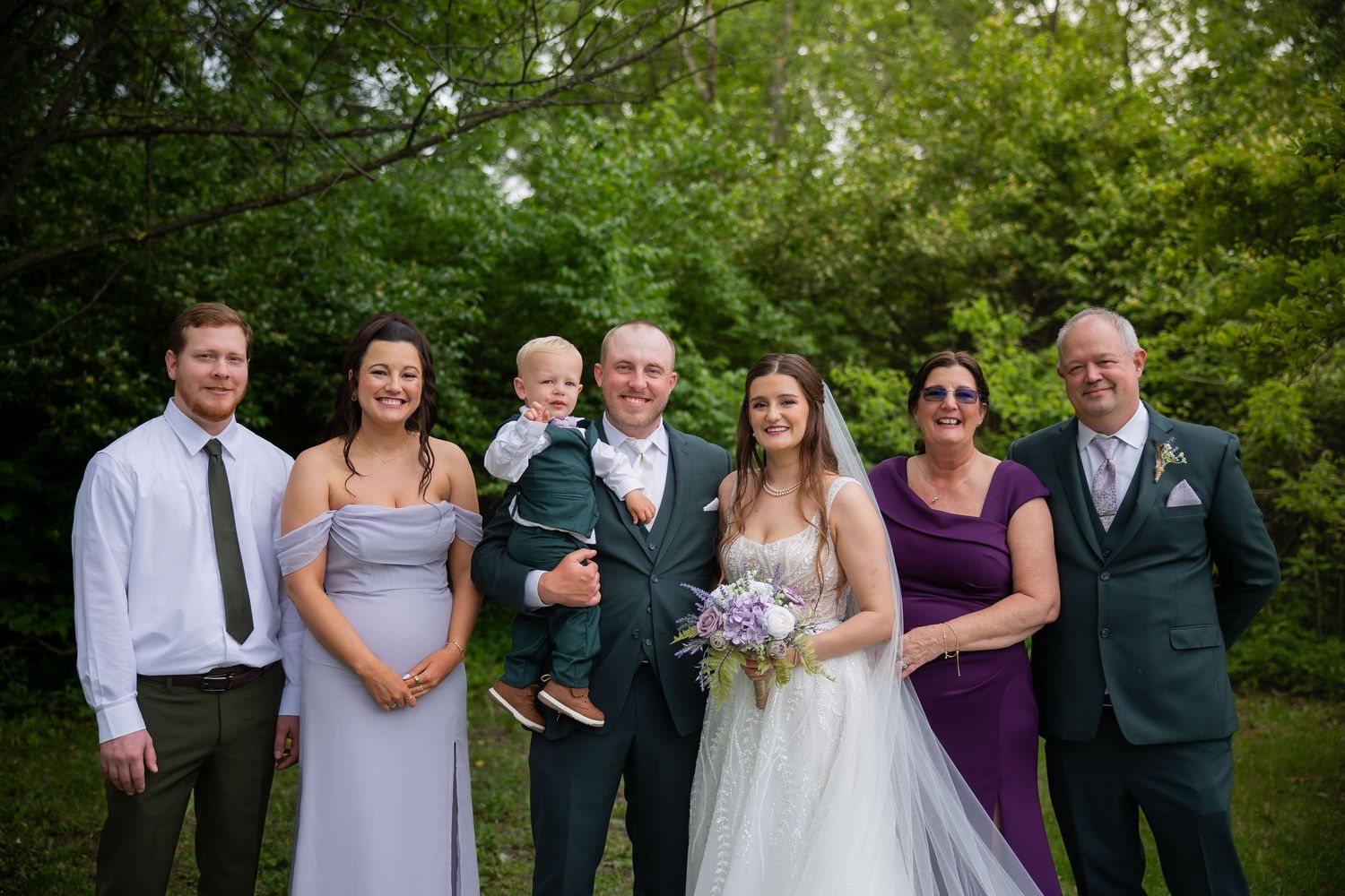 Wedding party group photo with bride, groom holding child, bridesmaids in lavender, and family in lush green setting.