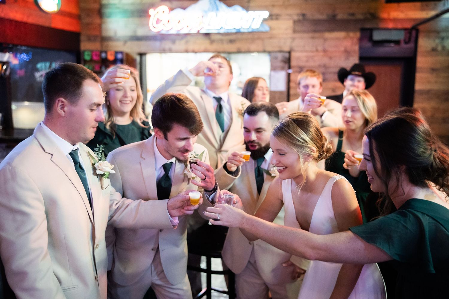 Wedding party toasts drinks together at a bar, bride in white gown surrounded by guests in formal attire.