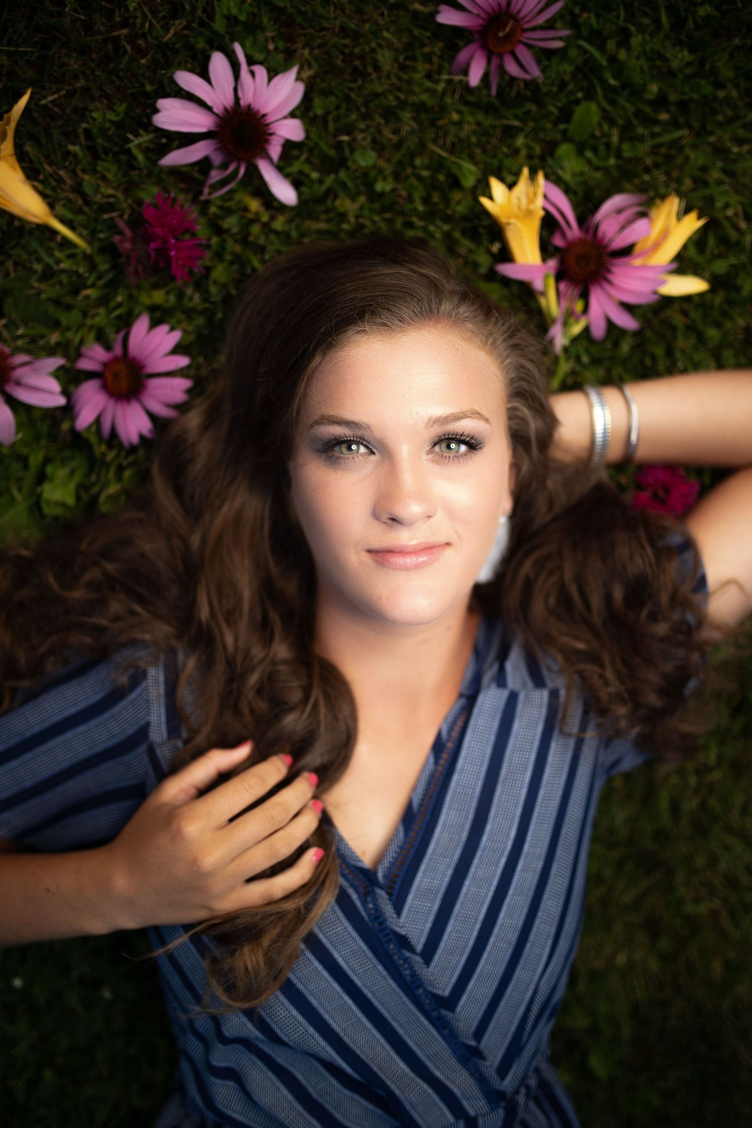 Young woman with long brown hair lying on green grass surrounded by colorful pink and yellow flowers, wearing a blue striped top.