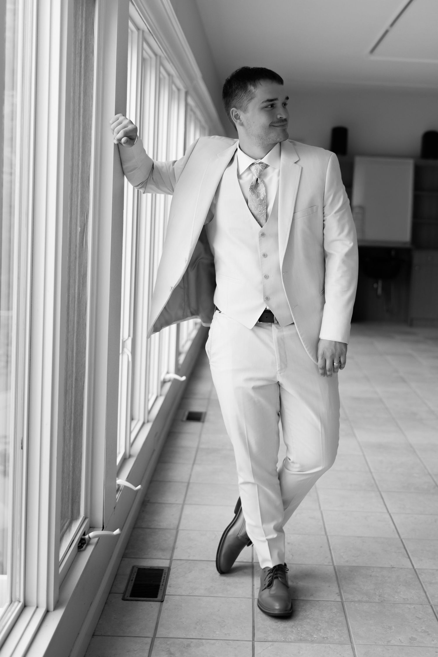 Groom in white suit leaning against windows in black and white wedding portrait.
