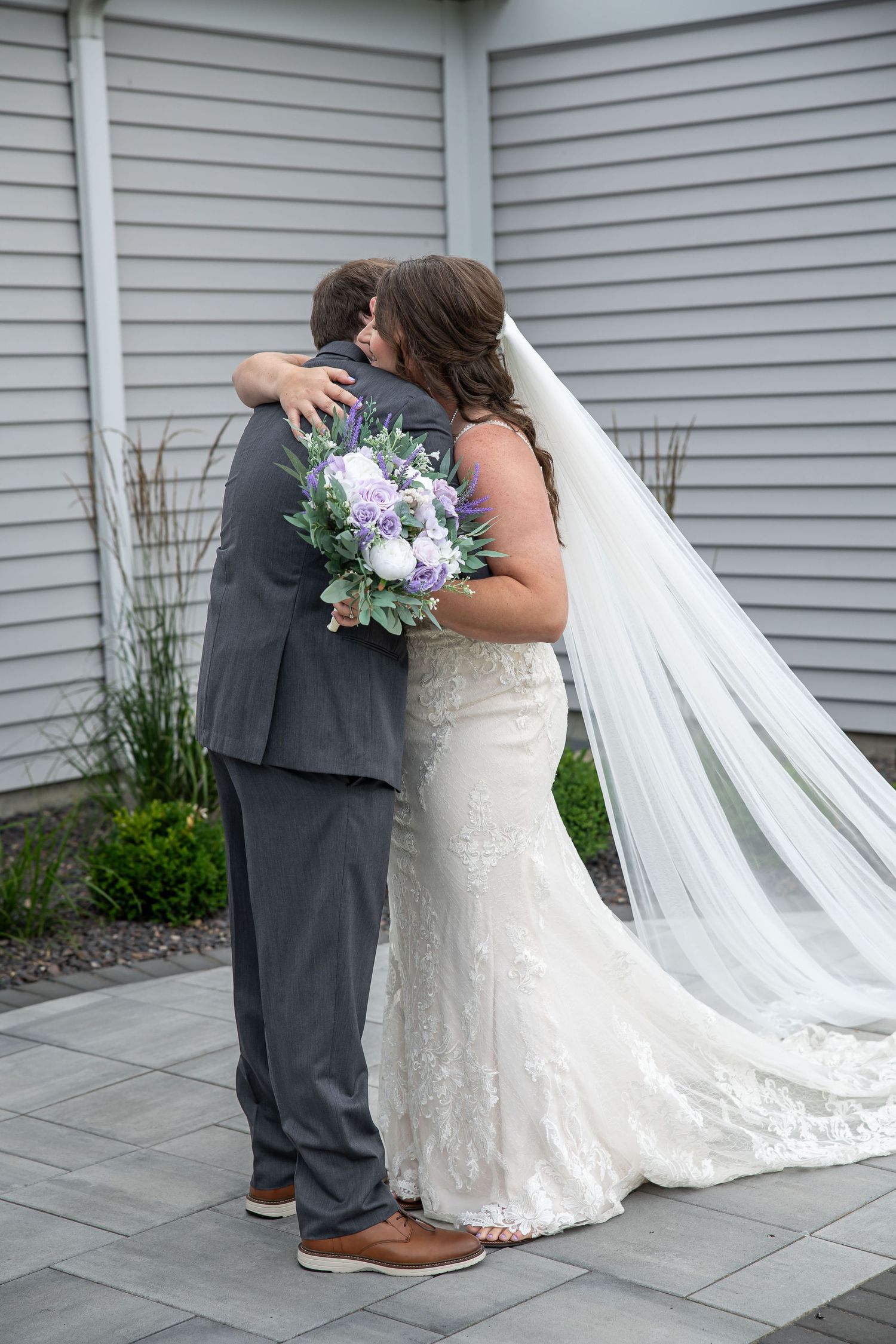Bride and groom sharing emotional first look hug, bride holding purple bouquet with flowing veil.