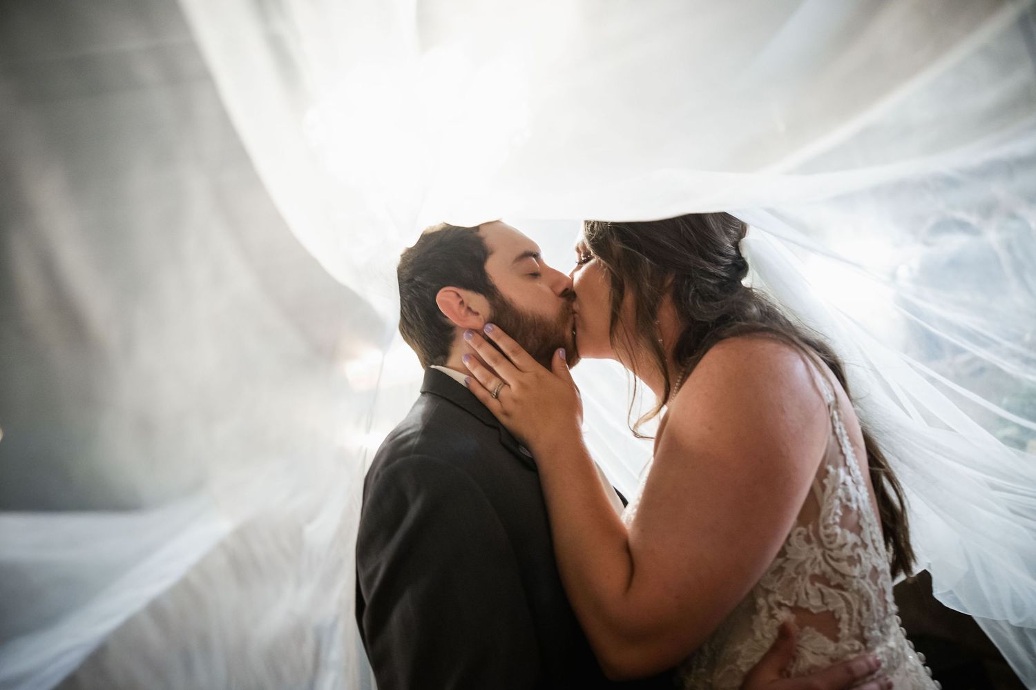 Bride and groom share a romantic kiss under a flowing white veil with soft backlight on their wedding day.