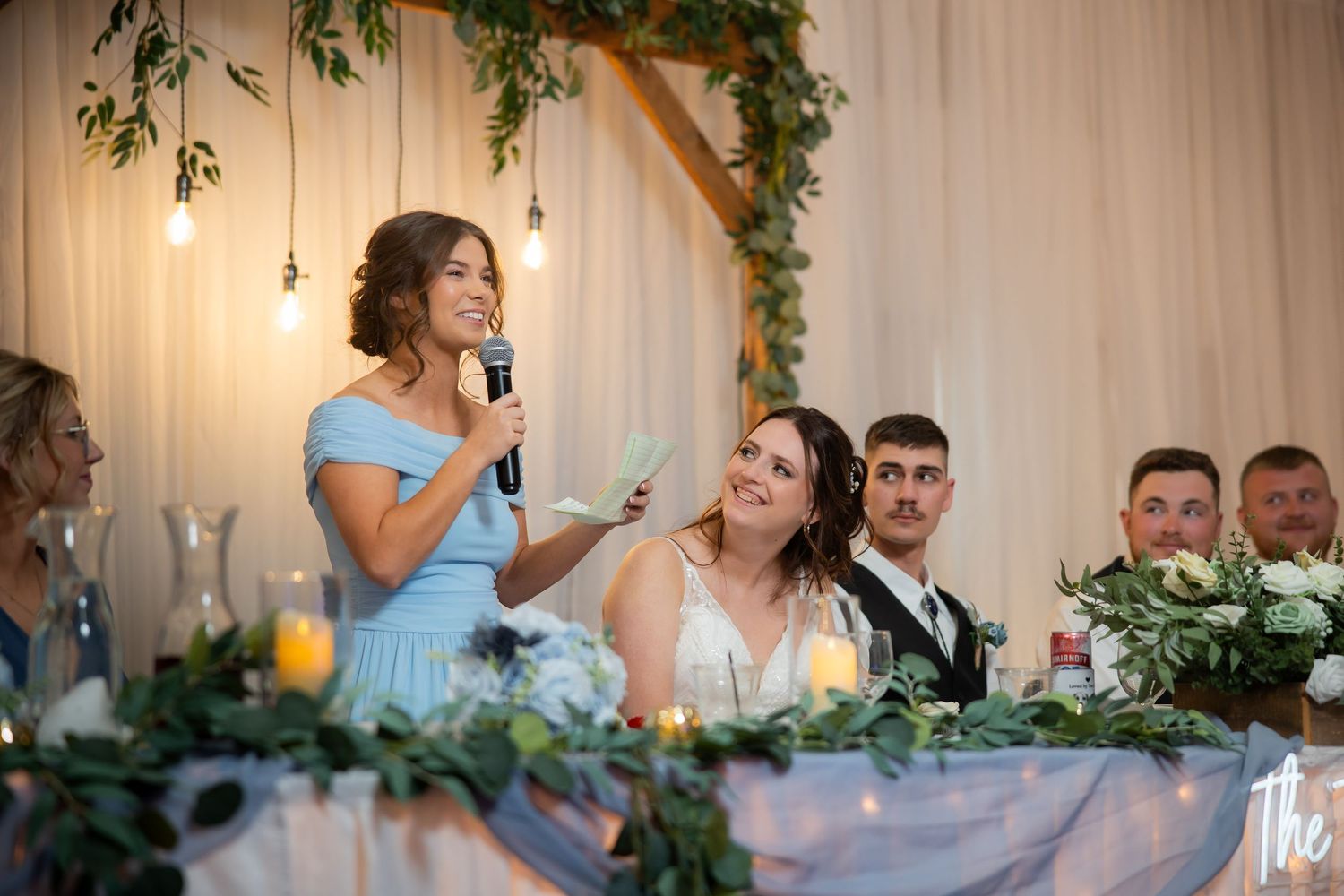 Bridesmaid in blue dress gives toast speech while smiling bride sits at floral decorated head table.