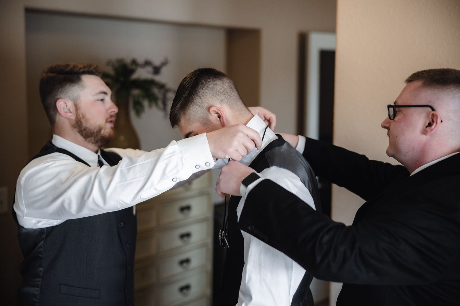Two groomsmen help a groom adjust his tie and suit jacket while getting ready for a wedding ceremony.