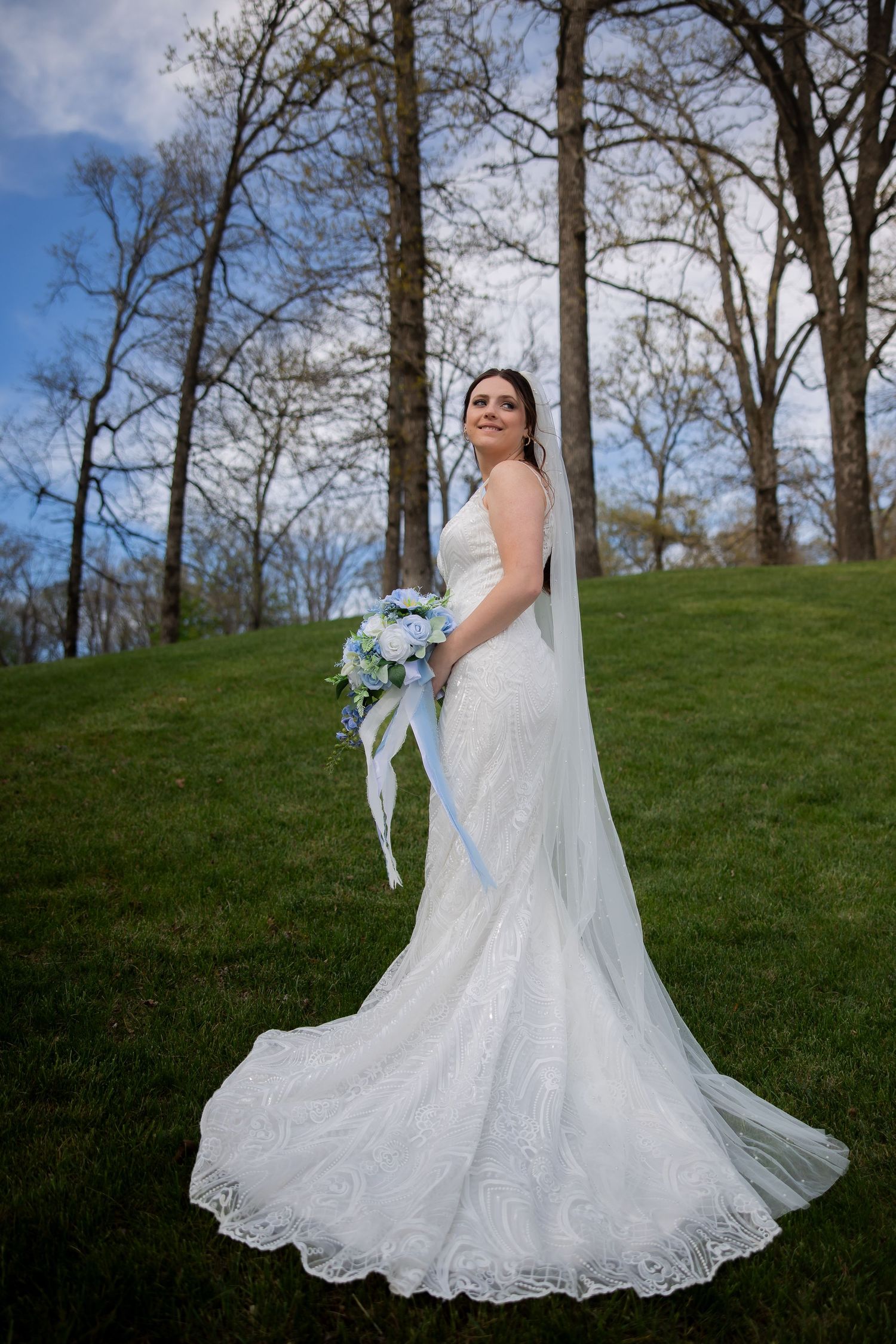 Bride in white lace mermaid gown with veil holds blue bouquet on green hillside with bare trees.