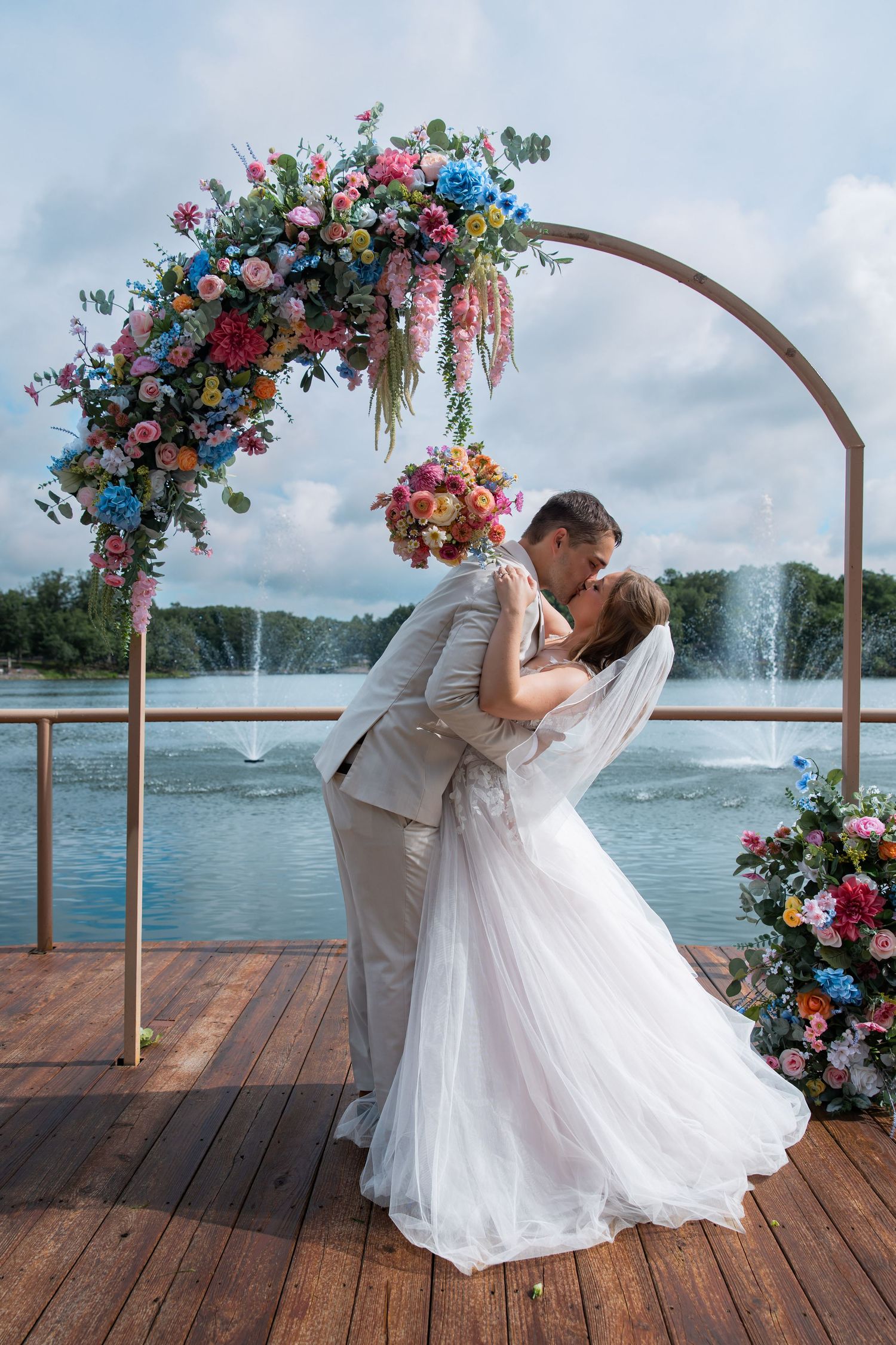 Bride and groom share a romantic kiss under a colorful floral arch on a lakeside deck with fountains in the background.