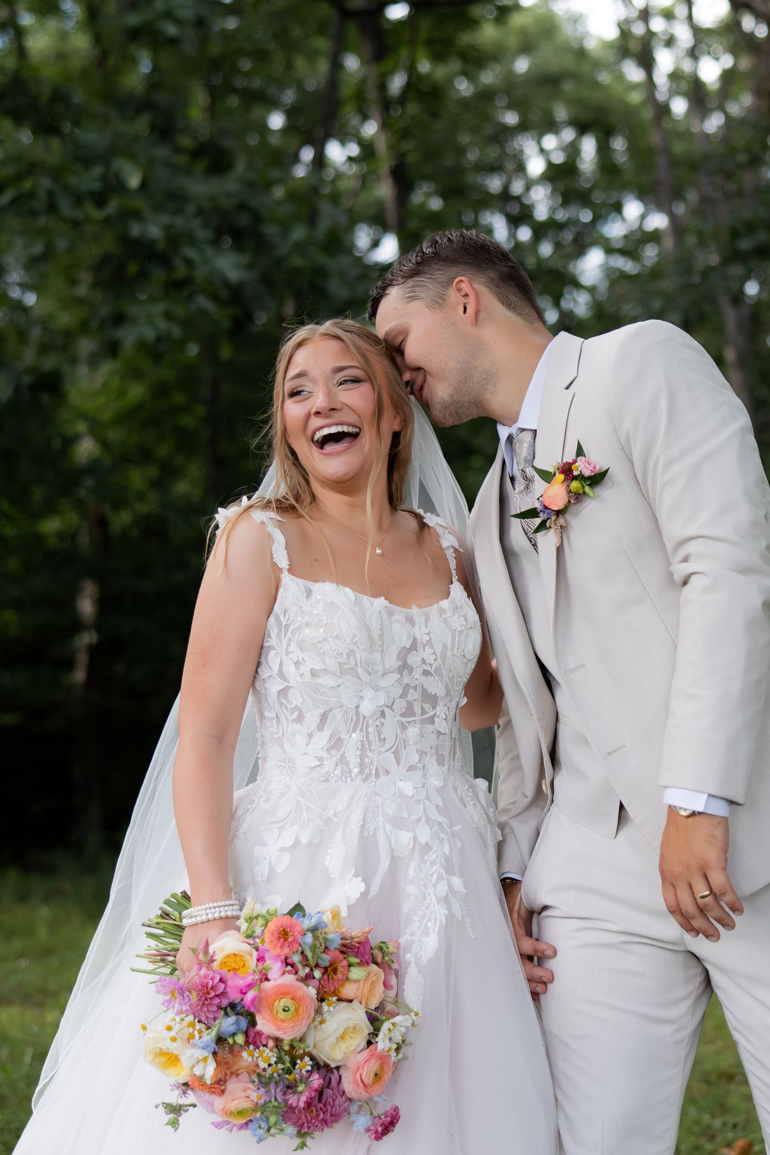 Joyful bride in white lace gown holds colorful bouquet as groom in beige suit whispers in her ear outdoors.