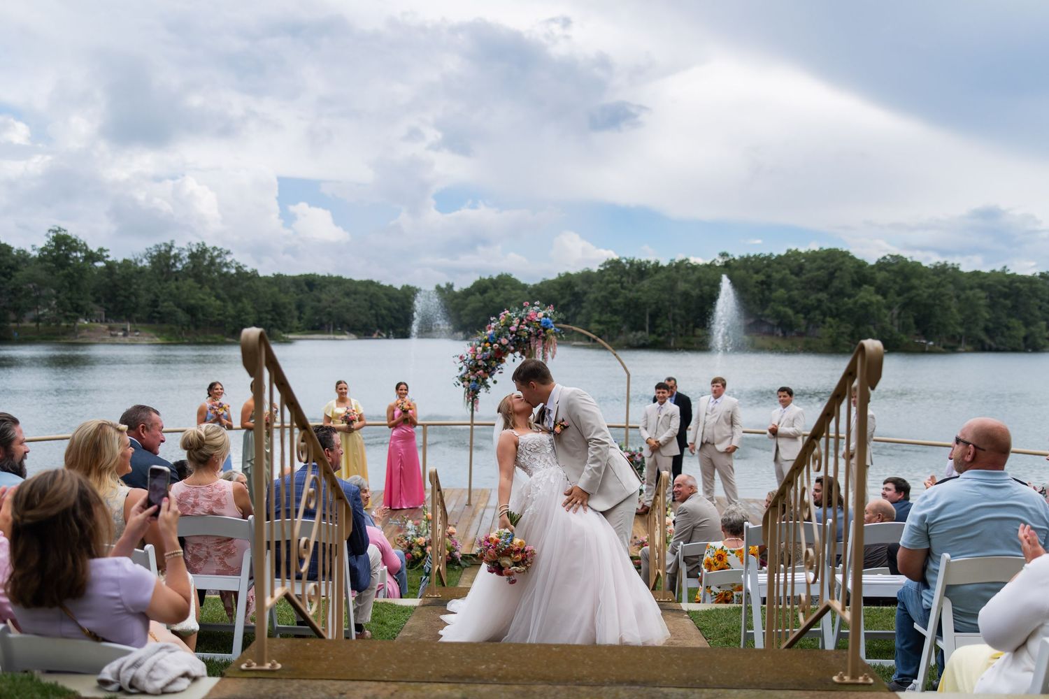 Couple shares first kiss at outdoor lakeside wedding ceremony with fountain backdrop.