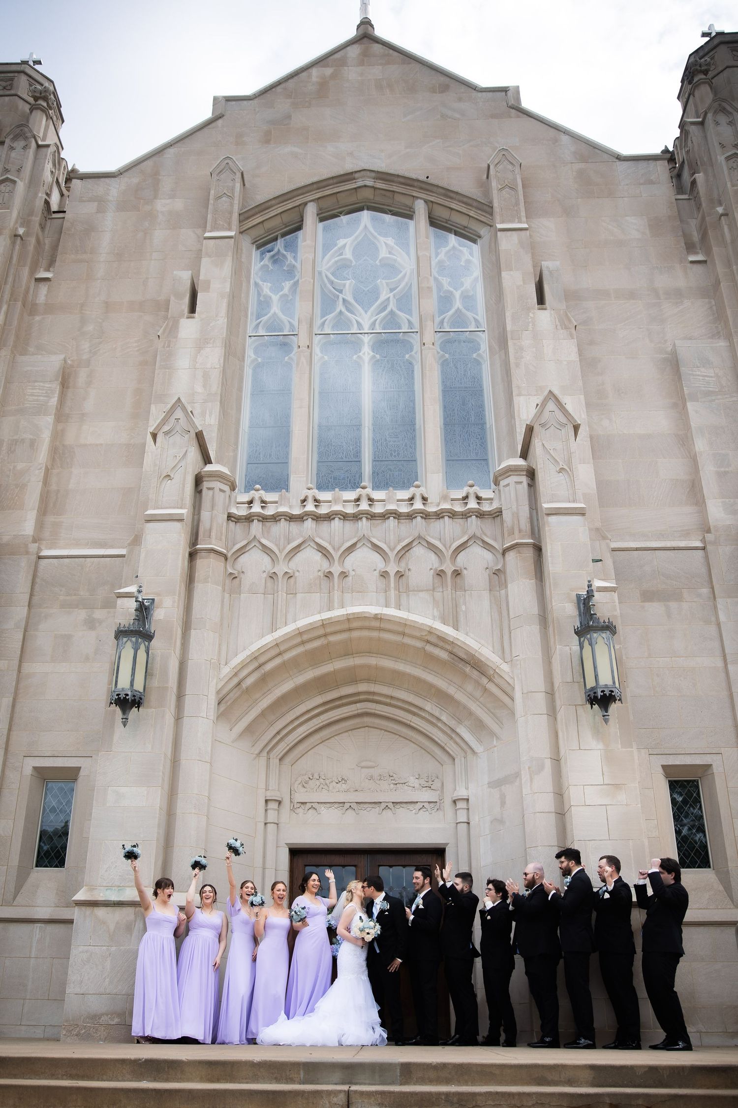 Wedding party poses on stone church steps with bridesmaids in lavender dresses.