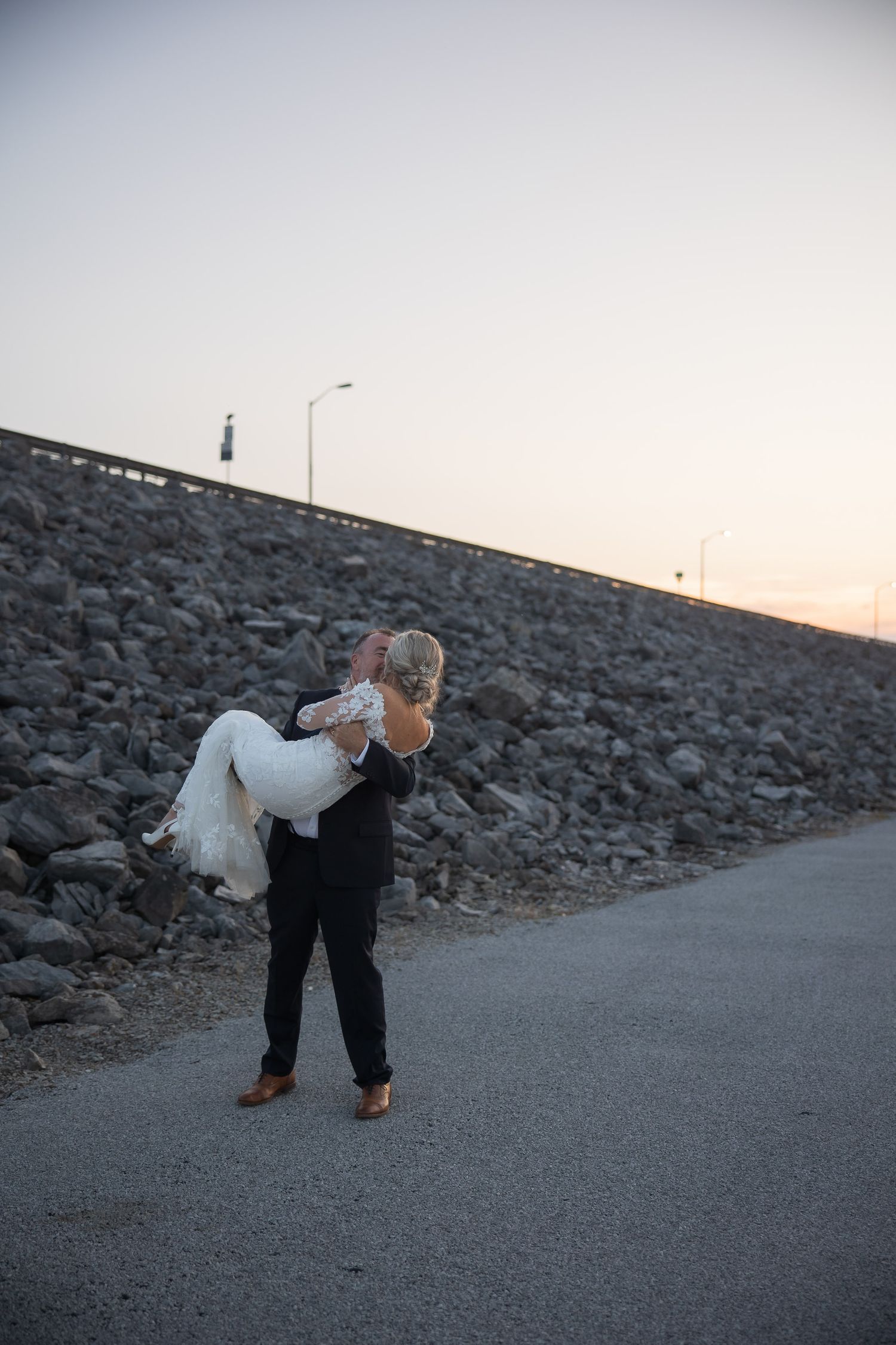A couple embraces on a rocky path at sunset with one person carrying the other in a romantic pose.