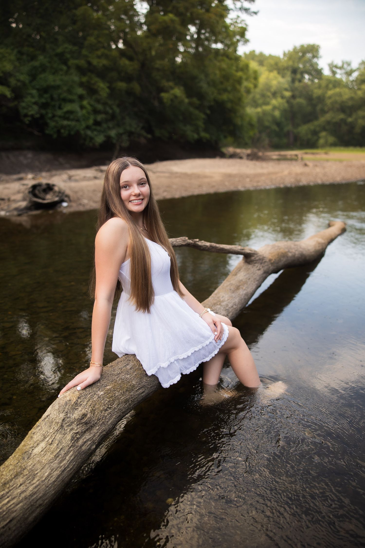 A person in a white sundress sits on a fallen log over a peaceful river in a natural outdoor setting.
