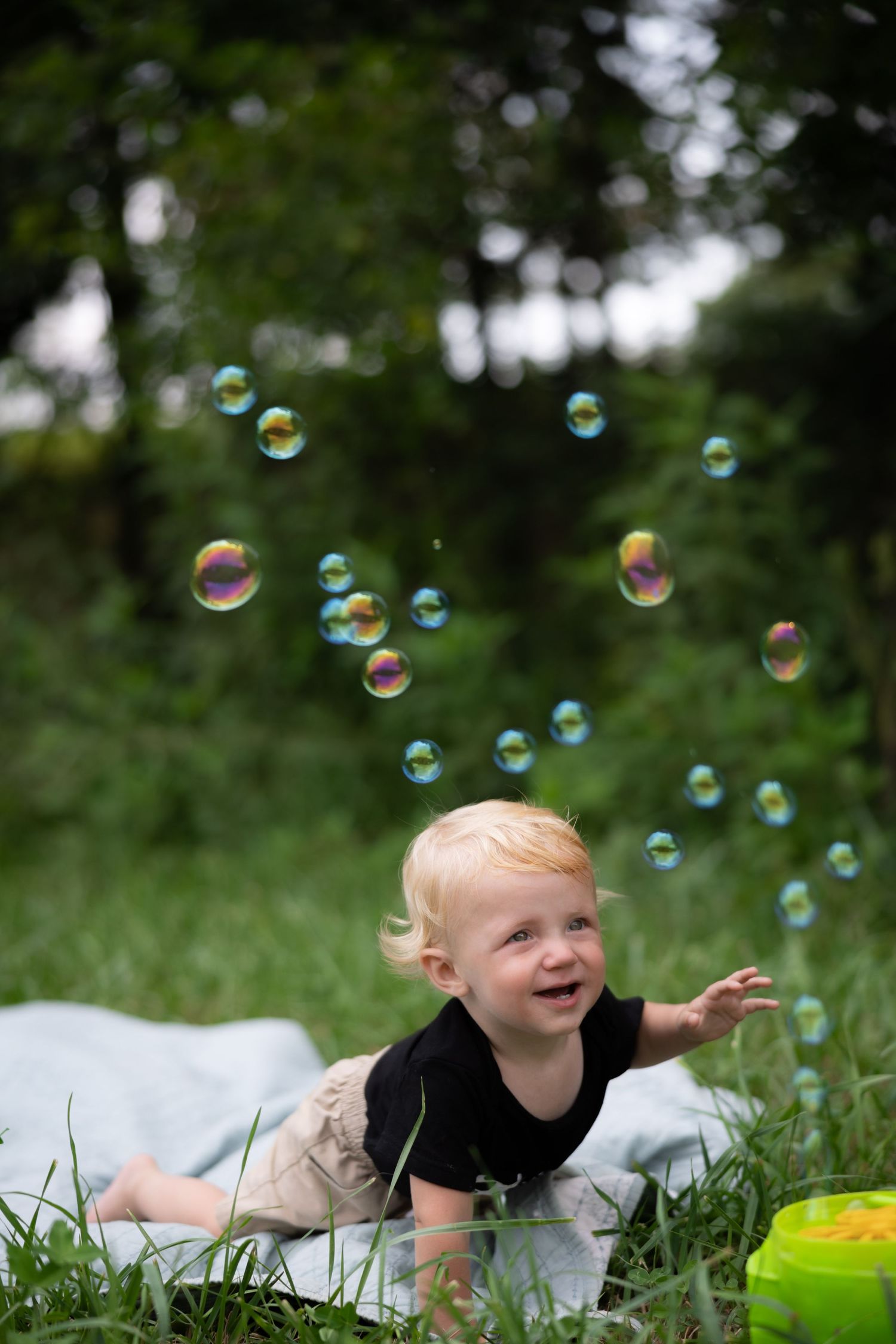Child lying on blanket playing with soap bubbles in green grass.
