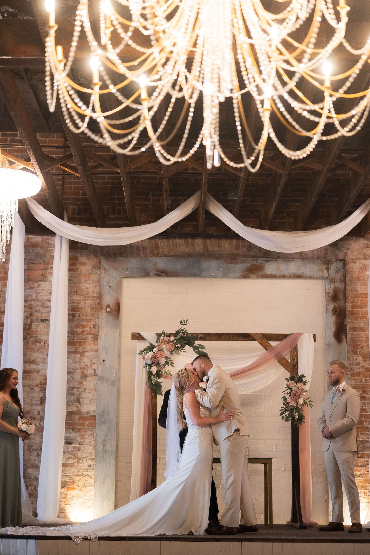Romantic wedding ceremony under elegant crystal chandelier and white draping in rustic brick venue.