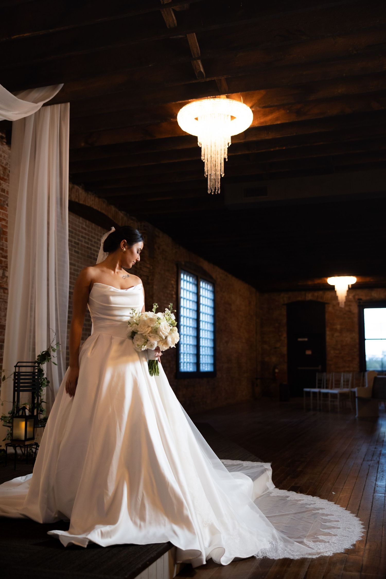 A stunning white wedding dress with a flowing train photographed in a rustic brick venue with elegant chandeliers.