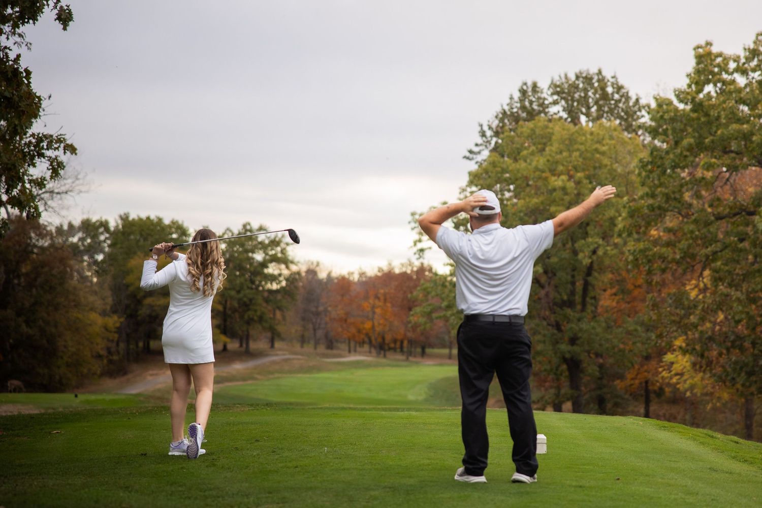 Golf course landscape with couple playfully posing on cart path surrounded by autumn trees.