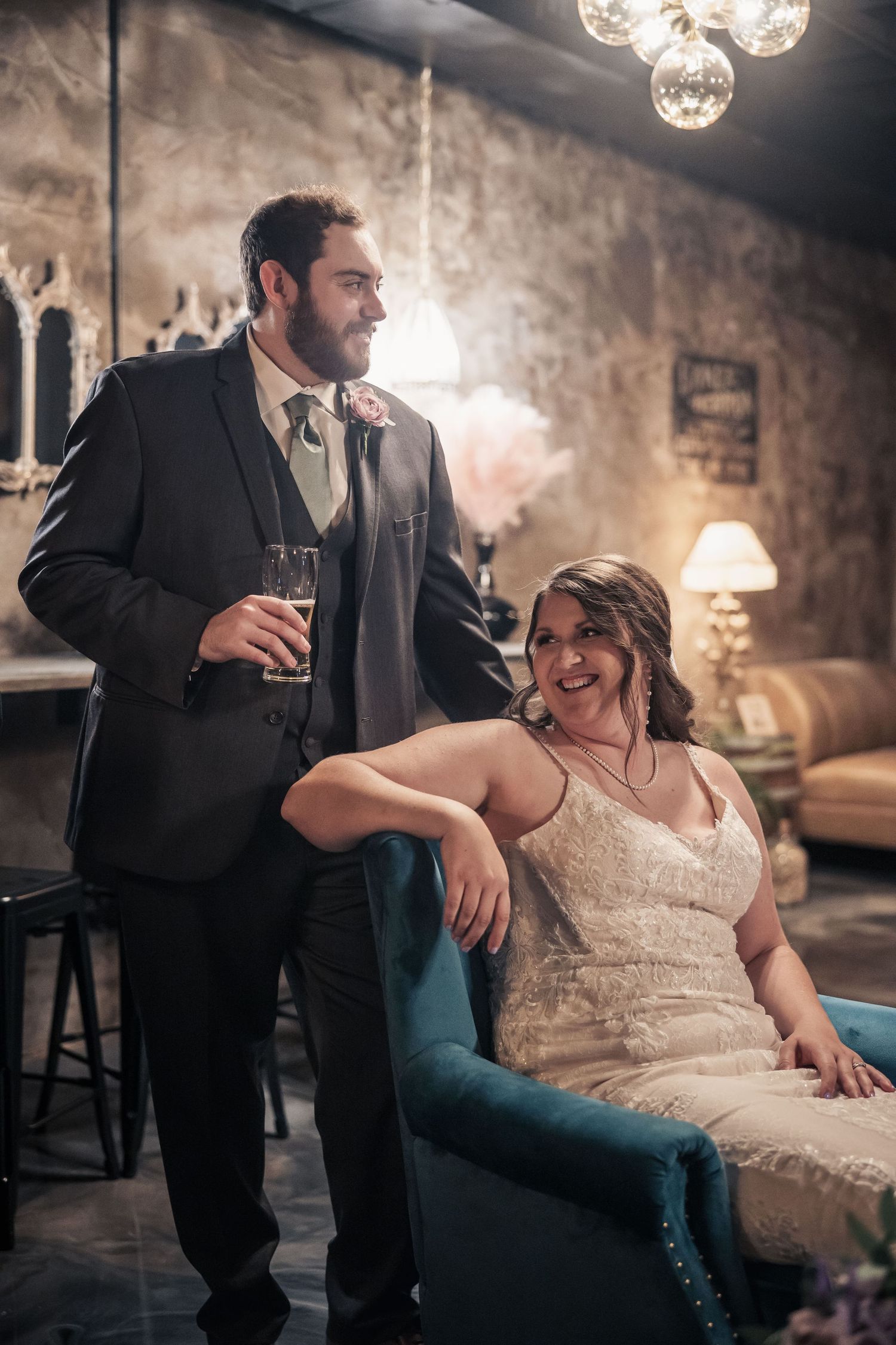 Bride in white gown sits in teal chair beside suited groom holding champagne in elegant, dimly lit wedding venue.