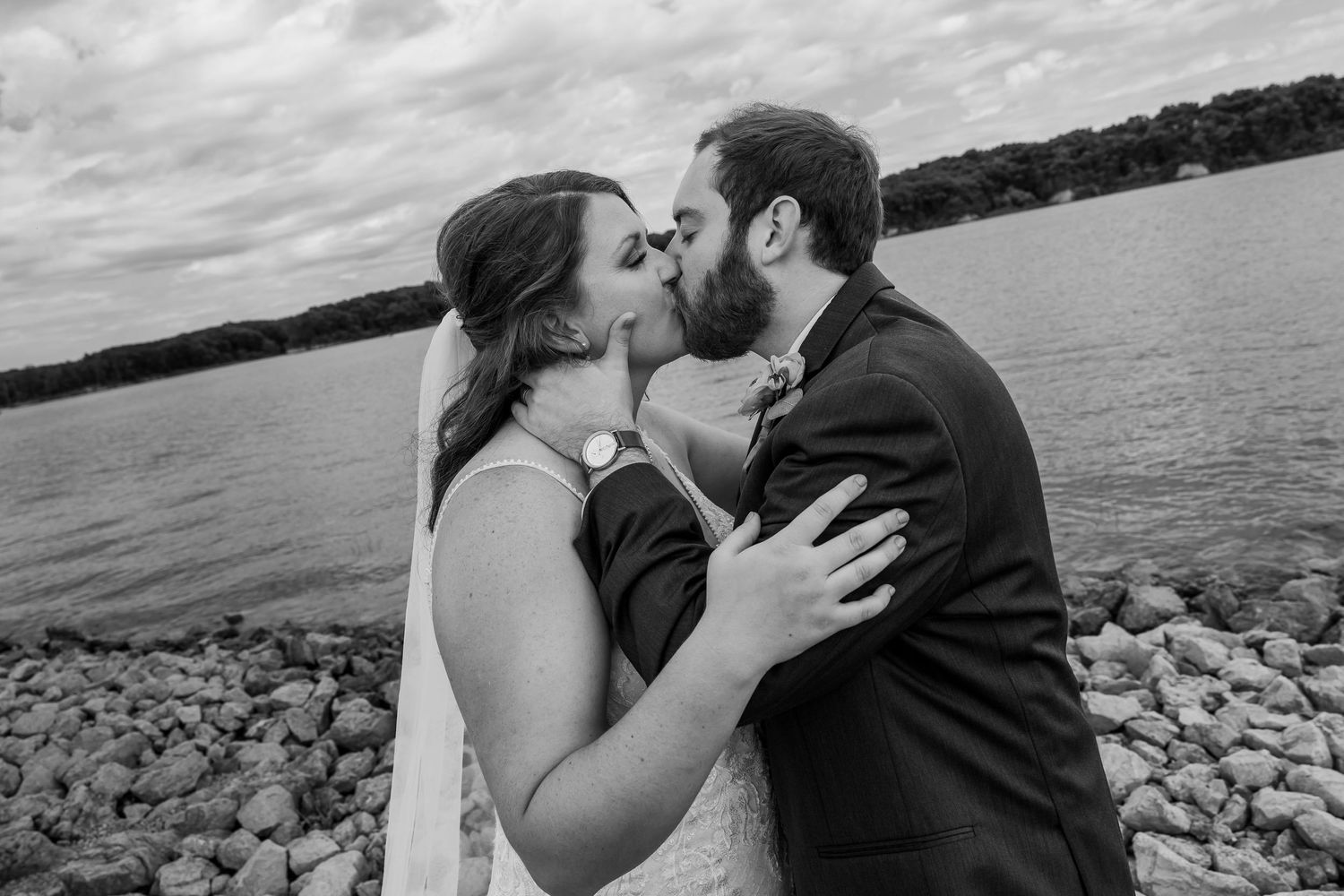 Black and white photo of a bride and groom sharing a kiss on a rocky shoreline with a lake and trees in the background.