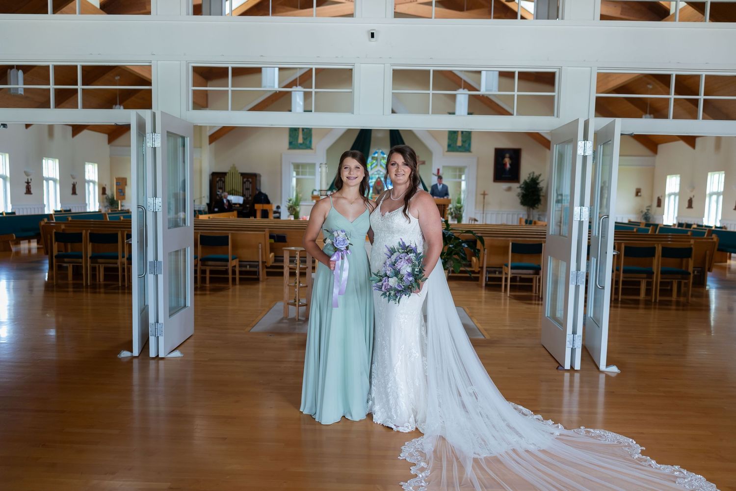 Bride in white gown and bridesmaid in sage green dress holding bouquets inside elegant wedding venue.