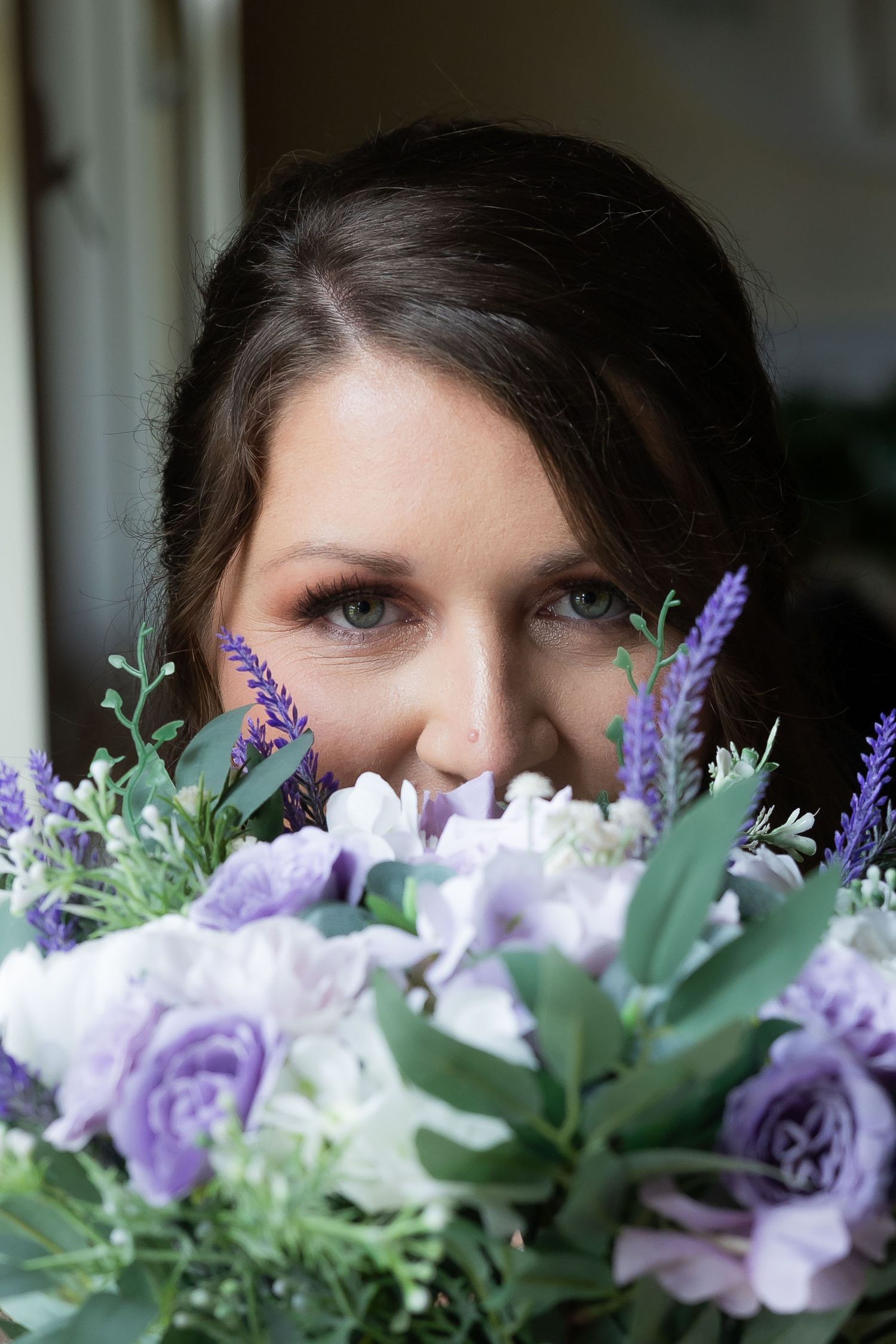 Close-up portrait of dark-haired bride peeking over purple and white wedding bouquet with greenery.