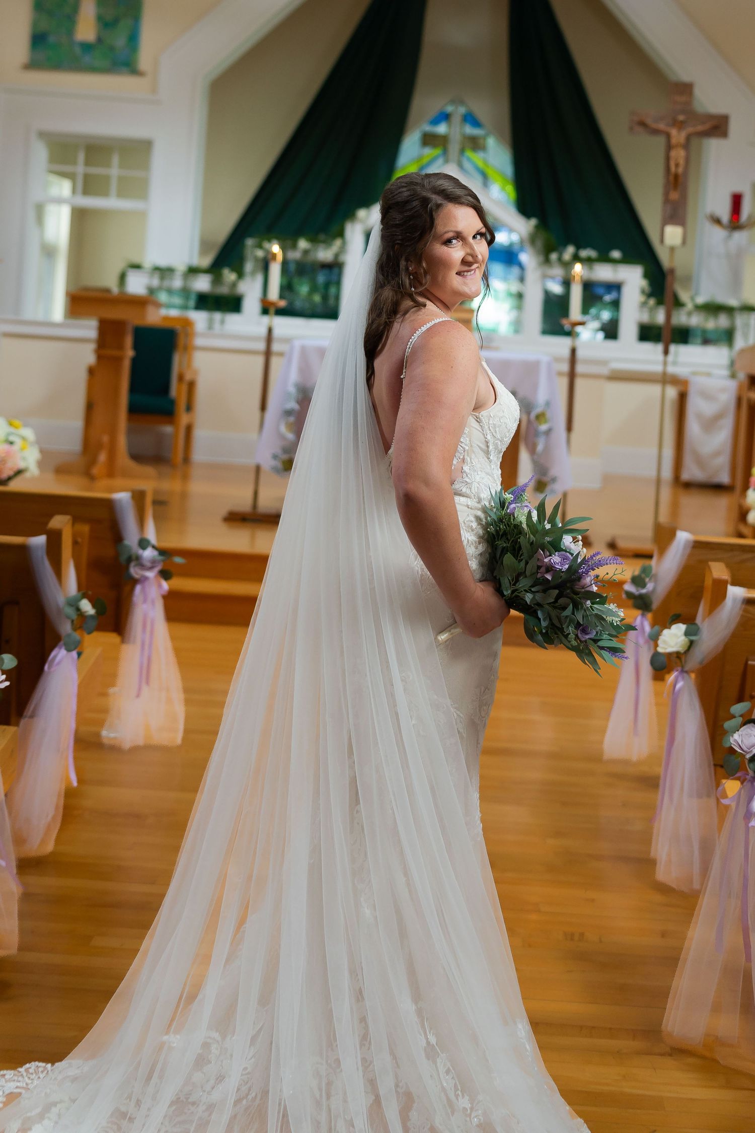 Bride in white lace gown and veil holds bouquet, posing in church aisle with pews and altar backdrop.