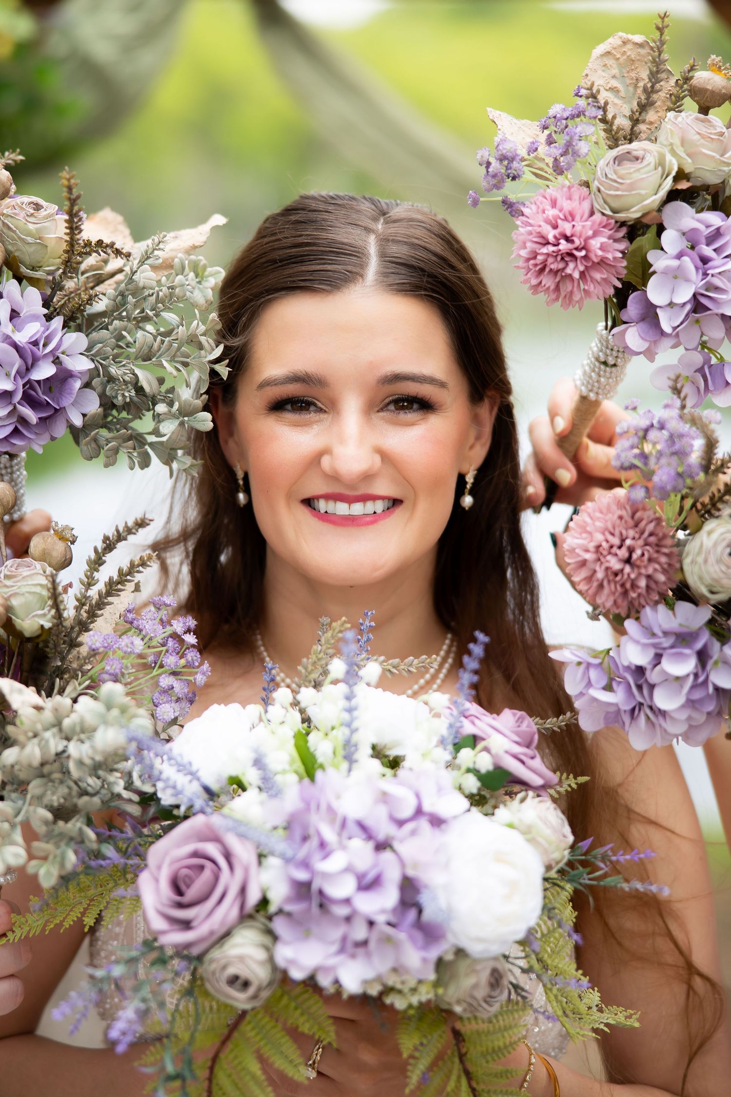 Smiling bride holding purple and white bouquet framed by floral wreaths with hydrangeas and roses outdoors.