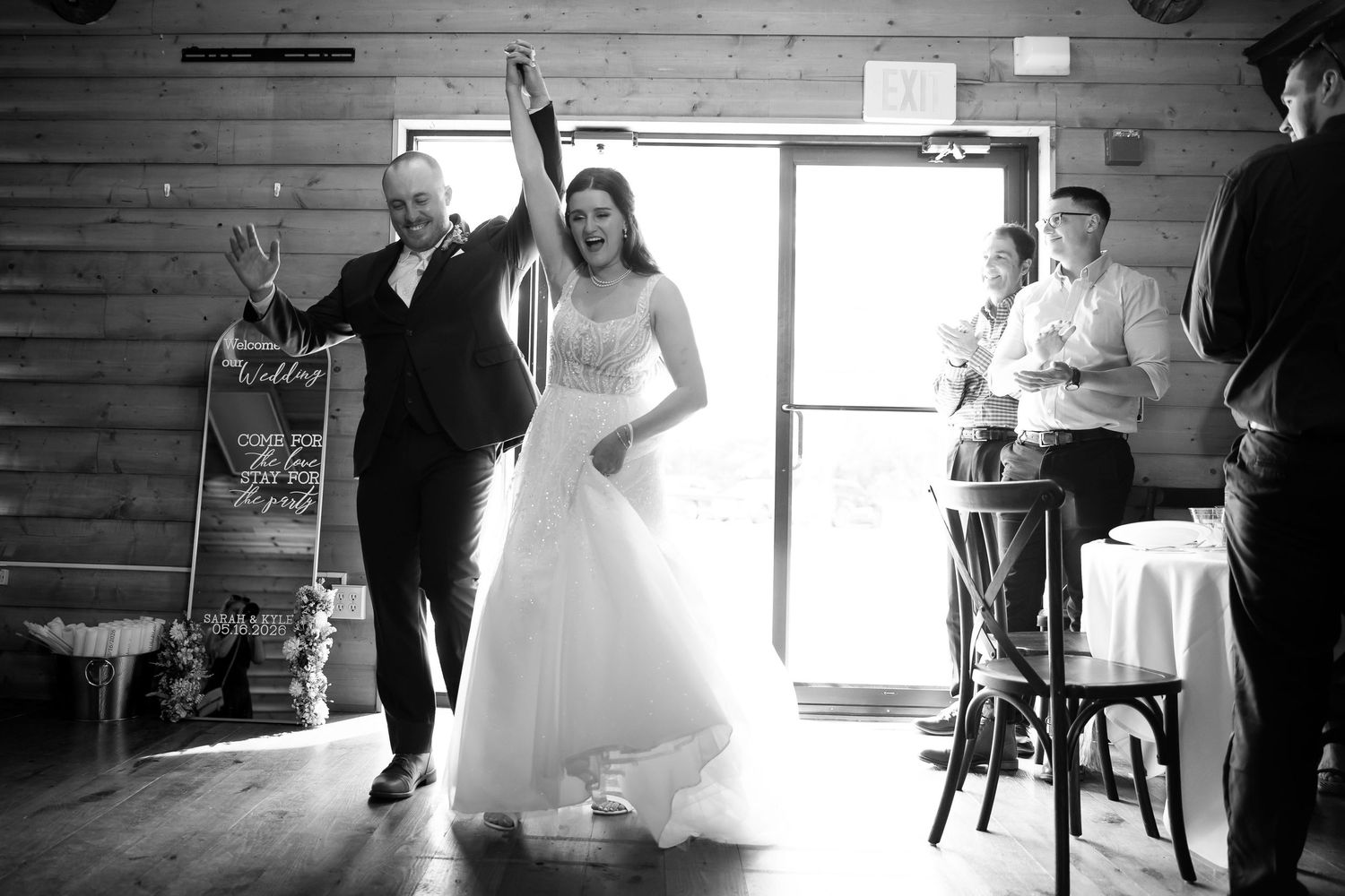 Bride and groom making a celebratory entrance into rustic wedding reception venue in black and white.