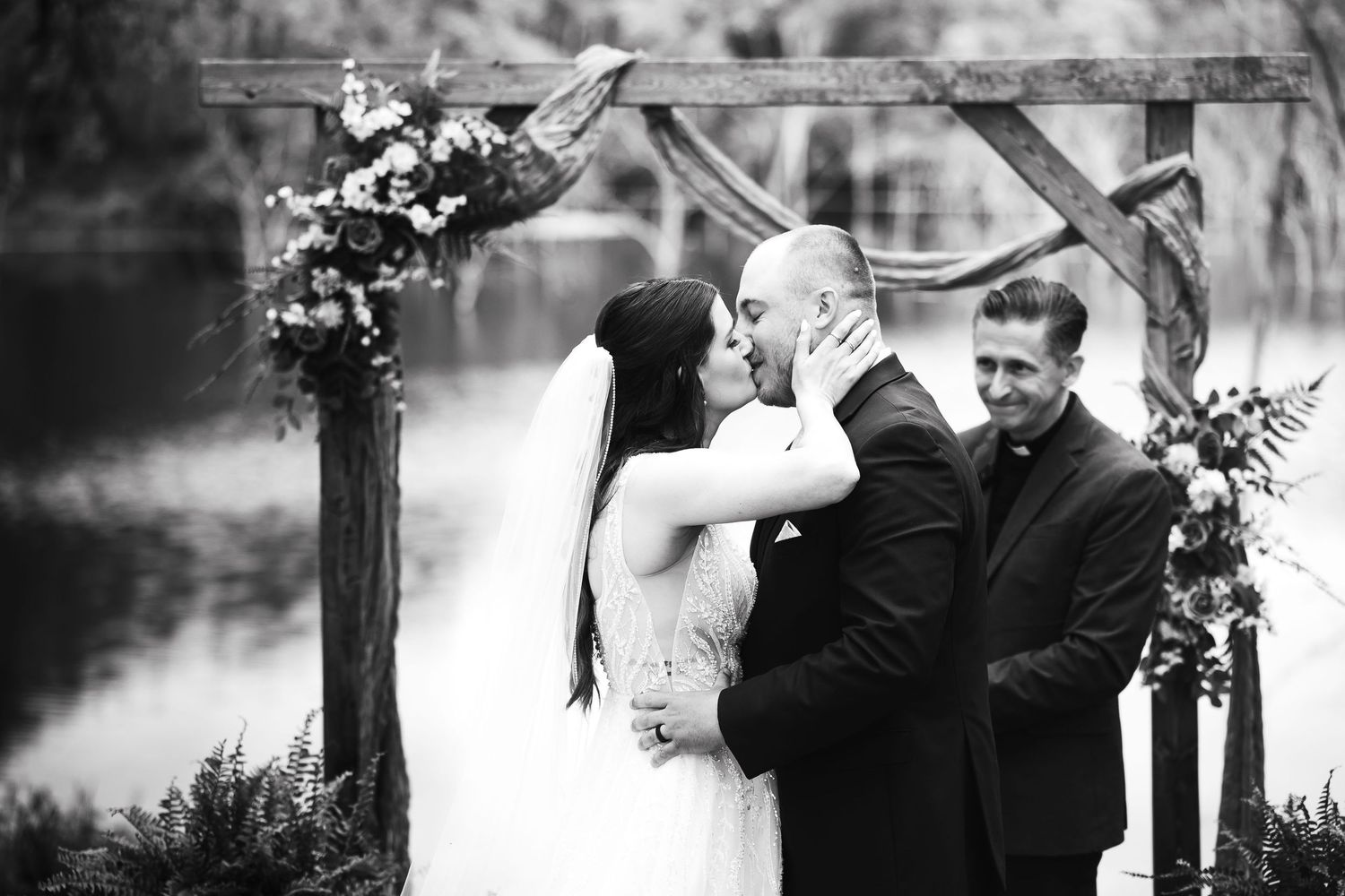 Bride and groom share first kiss at outdoor wedding ceremony with floral arch and officiant in black and white photo.
