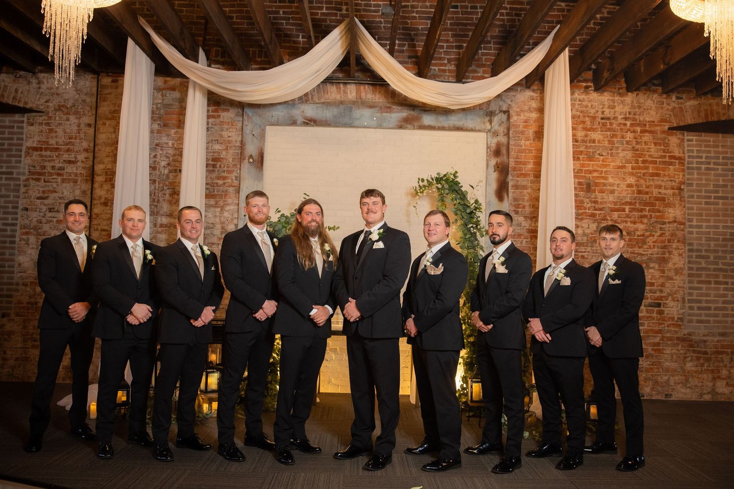 Wedding party members lined up against exposed brick wall under warm lighting.