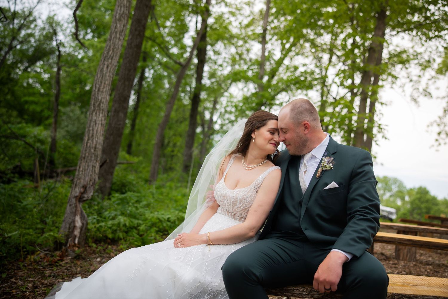 Romantic bride and groom share an intimate moment seated on a bench surrounded by tall trees in a woodland setting.