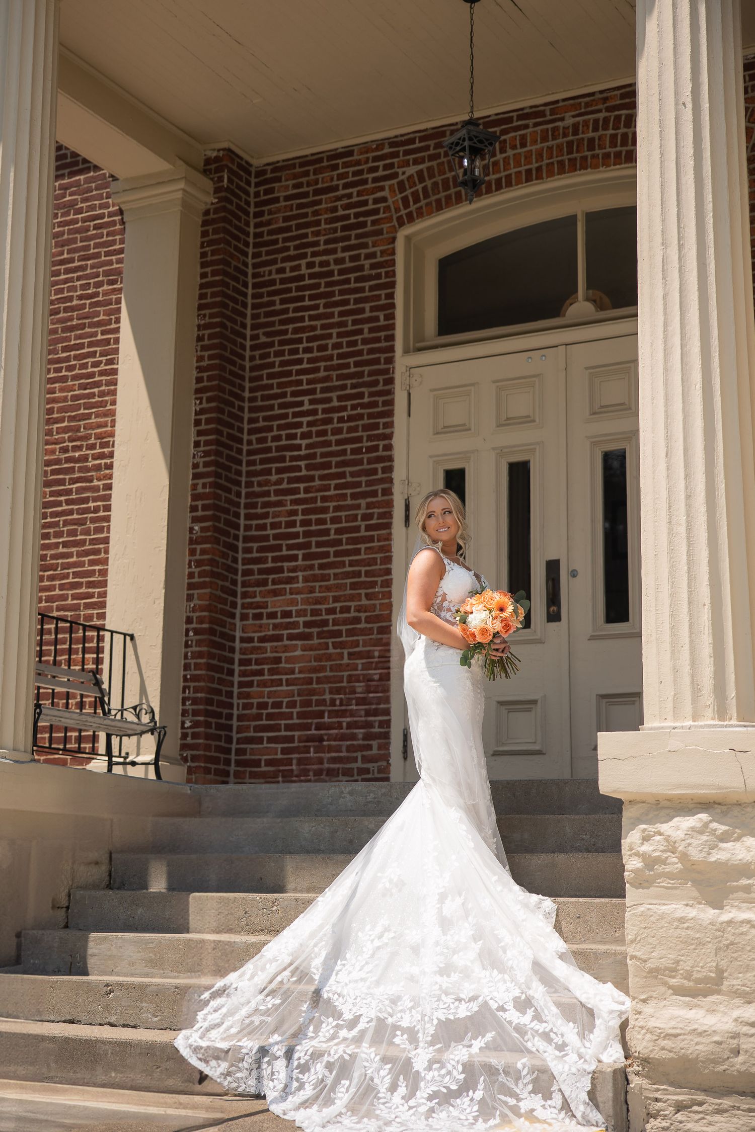 Stunning lace wedding dress with cathedral train photographed against historic brick building columns.