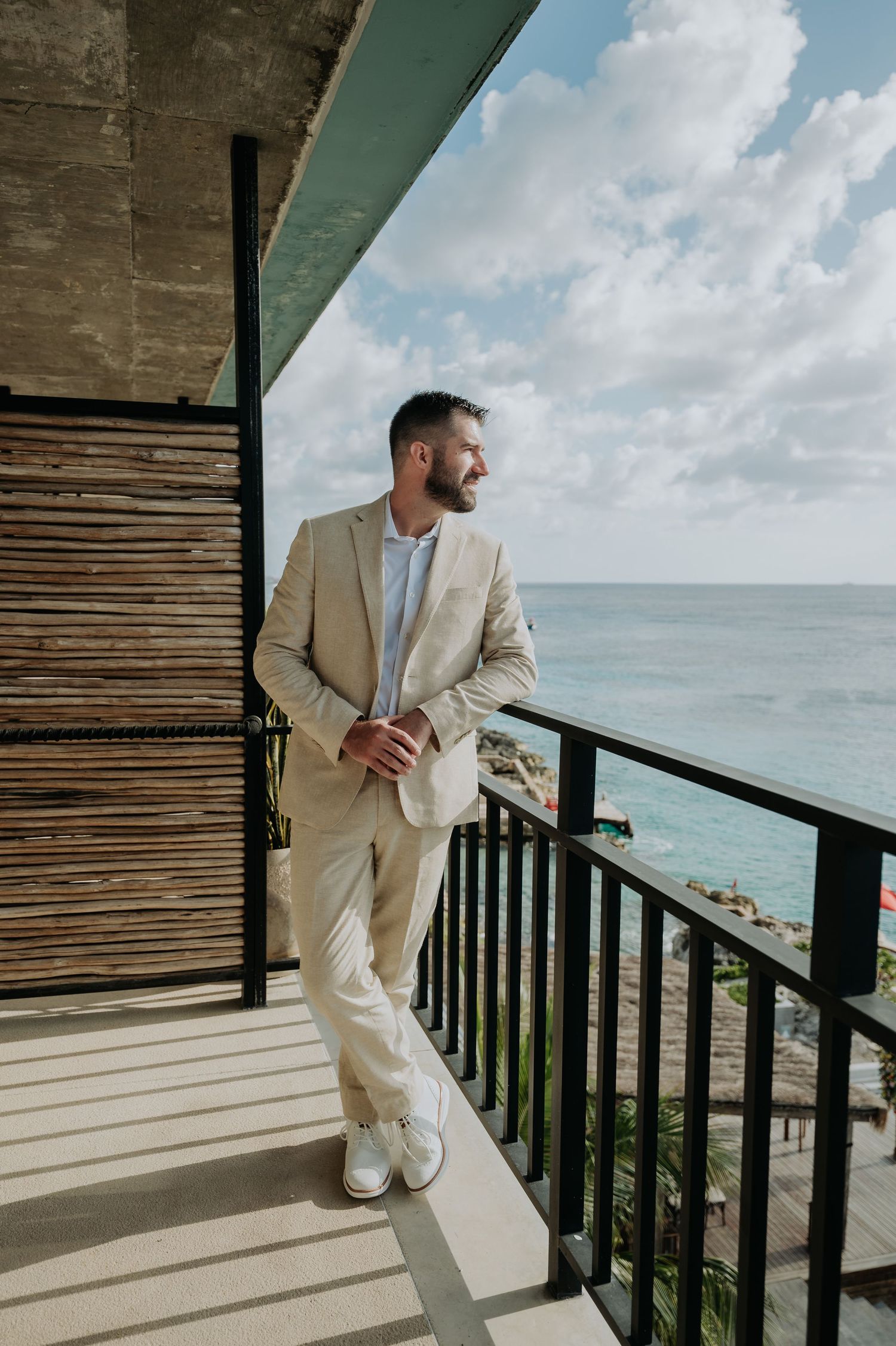 Sequence of a groom in a light colored suit posing on a wooden balcony overlooking the ocean.