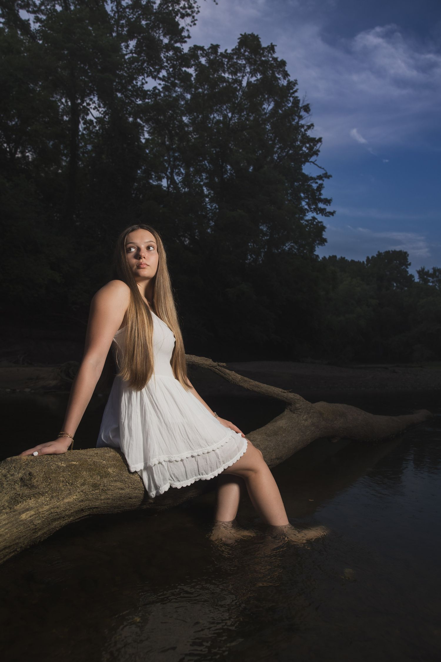 A person in a white dress sits on a log at dusk with feet dangling in dark water.