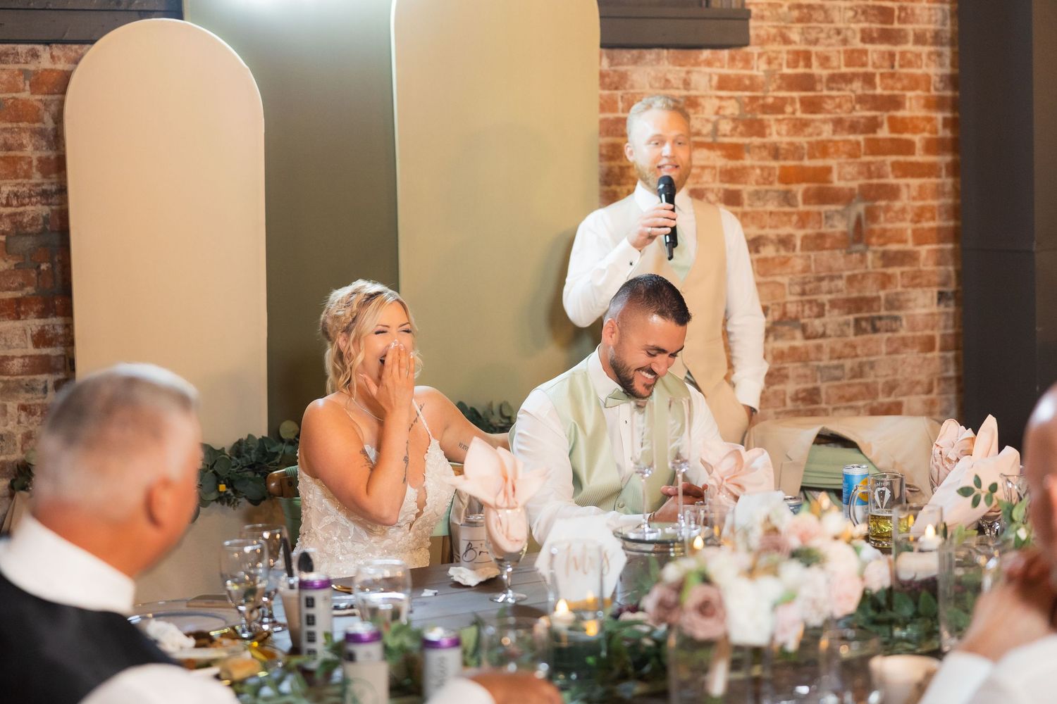 Candid reception moment during wedding speeches in industrial venue with brick walls.