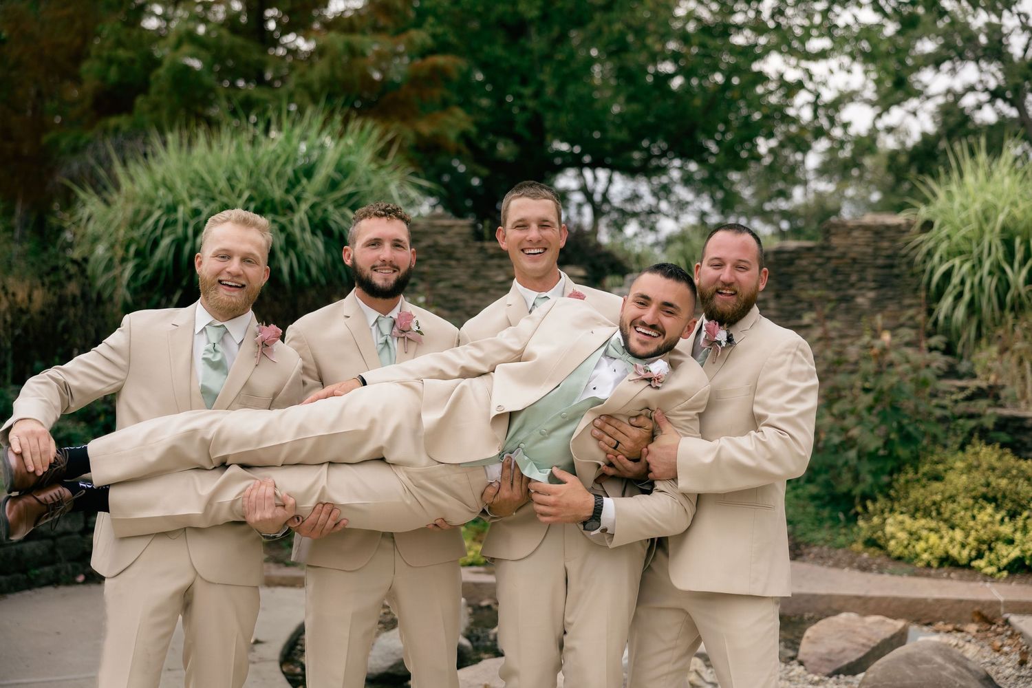 Groomsmen in beige suits share a candid moment while sitting and laughing together on stone steps outdoors.