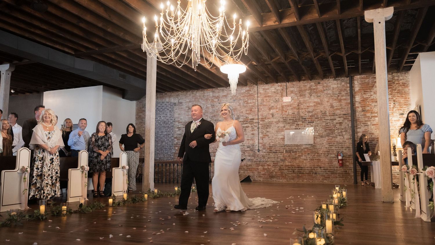 Wedding celebration in industrial venue with chandelier and candlelit aisle.