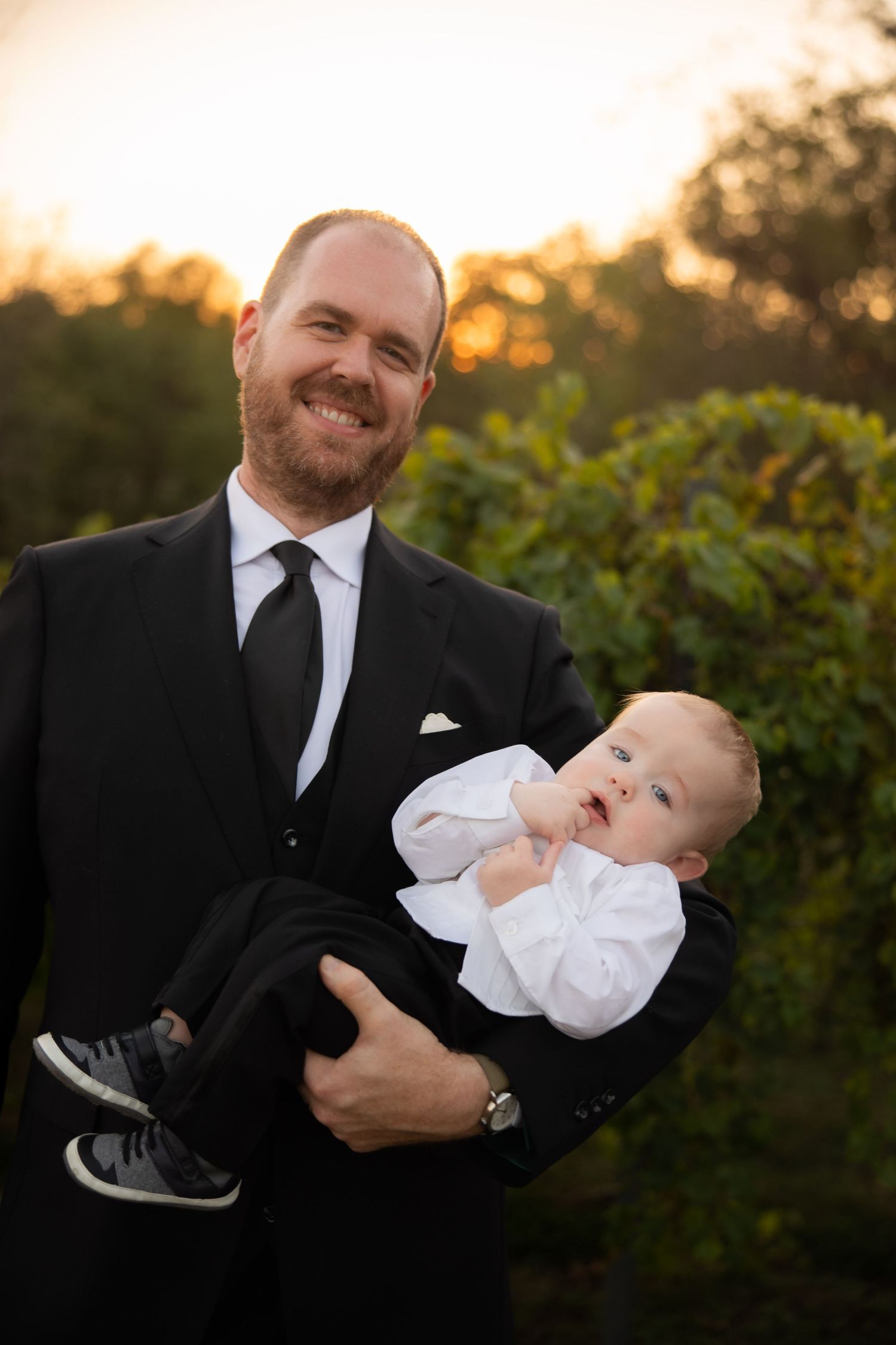 Joyful moments between a man in formal attire and baby in white clothing at sunset.