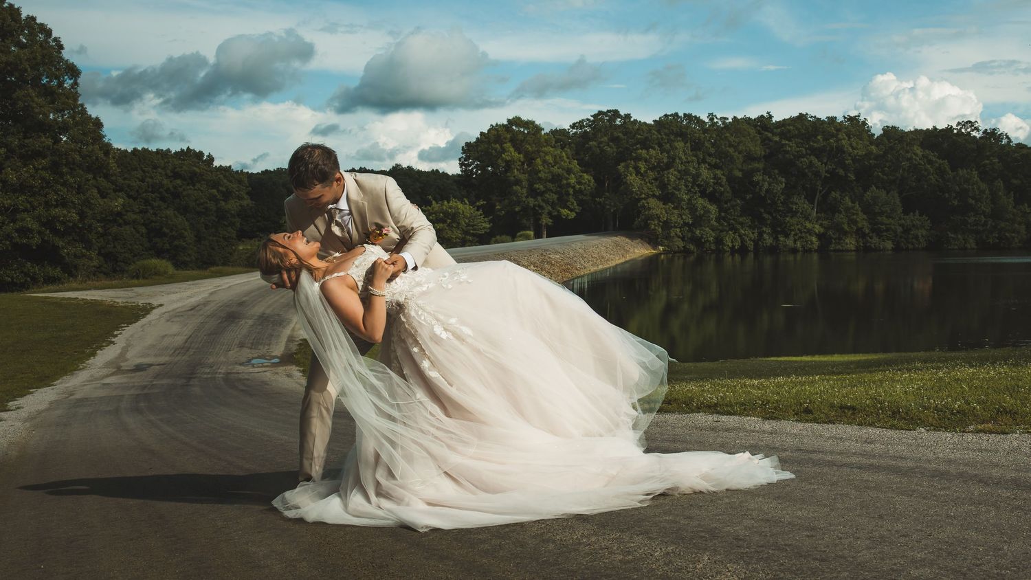 A groom dips his bride in a romantic wedding photo by a serene lake surrounded by lush green trees.