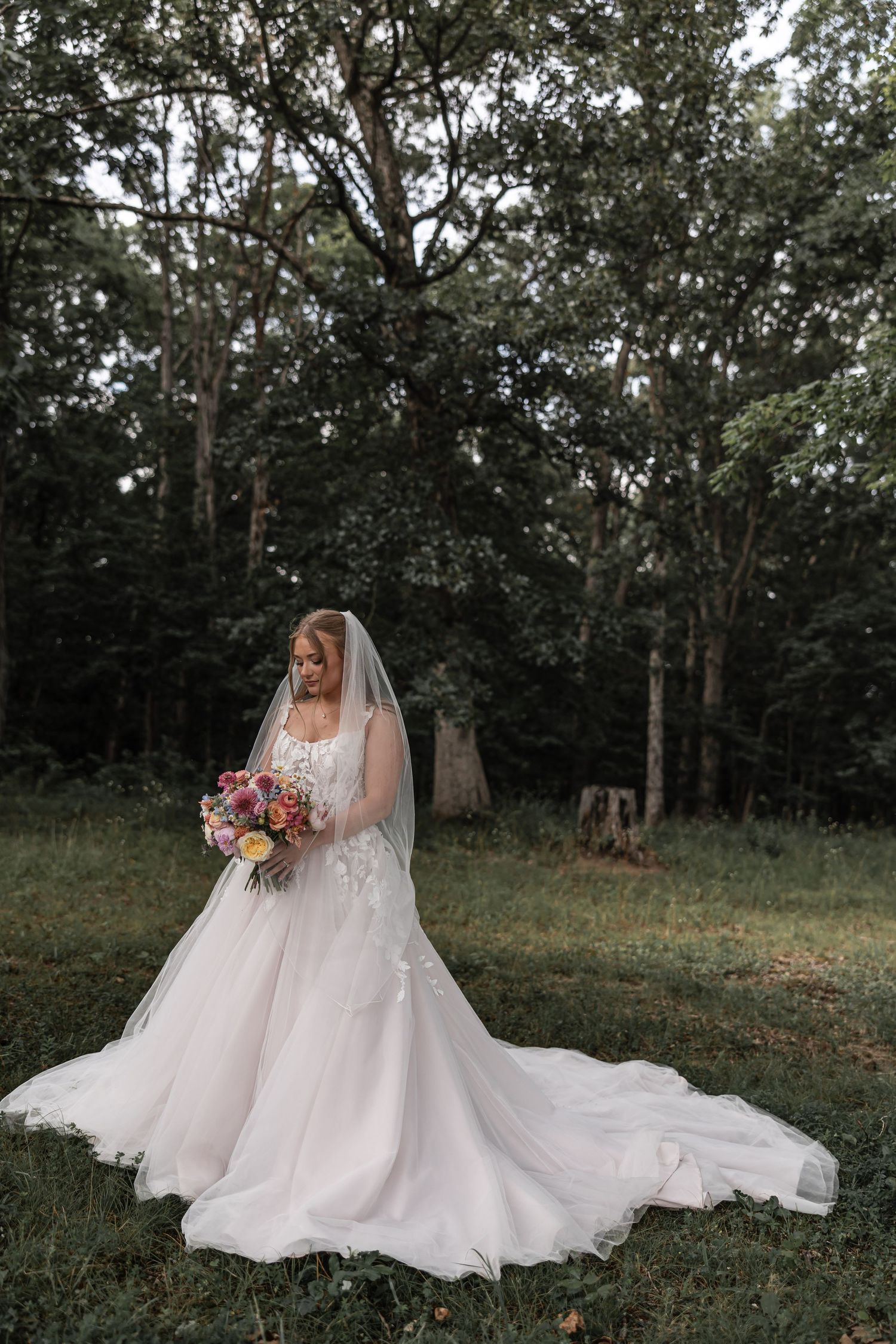 Elegant bride in a flowing white gown with veil holding a colorful bouquet in a lush green forest setting.